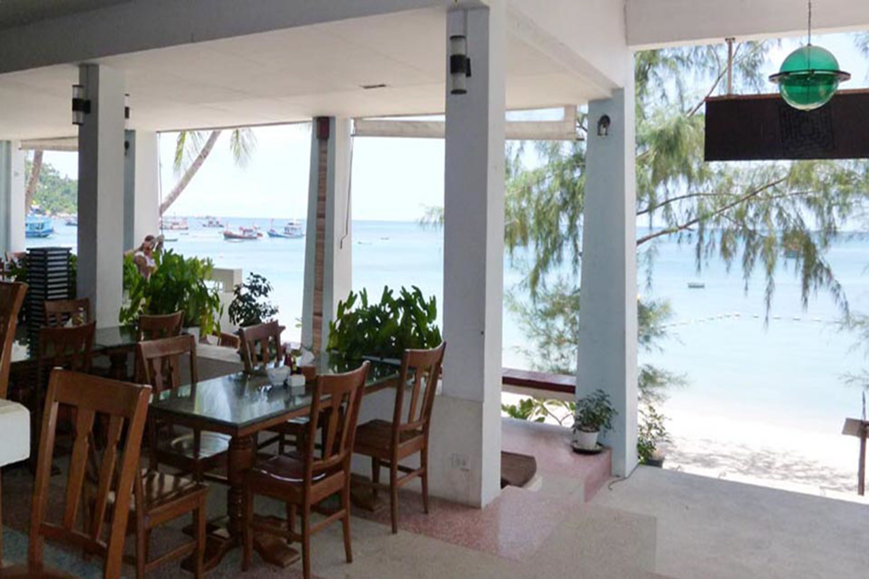 Un coin repas avec une table en verre et des chaises en bois, donnant sur une plage où l’on aperçoit des bateaux et des palmiers.