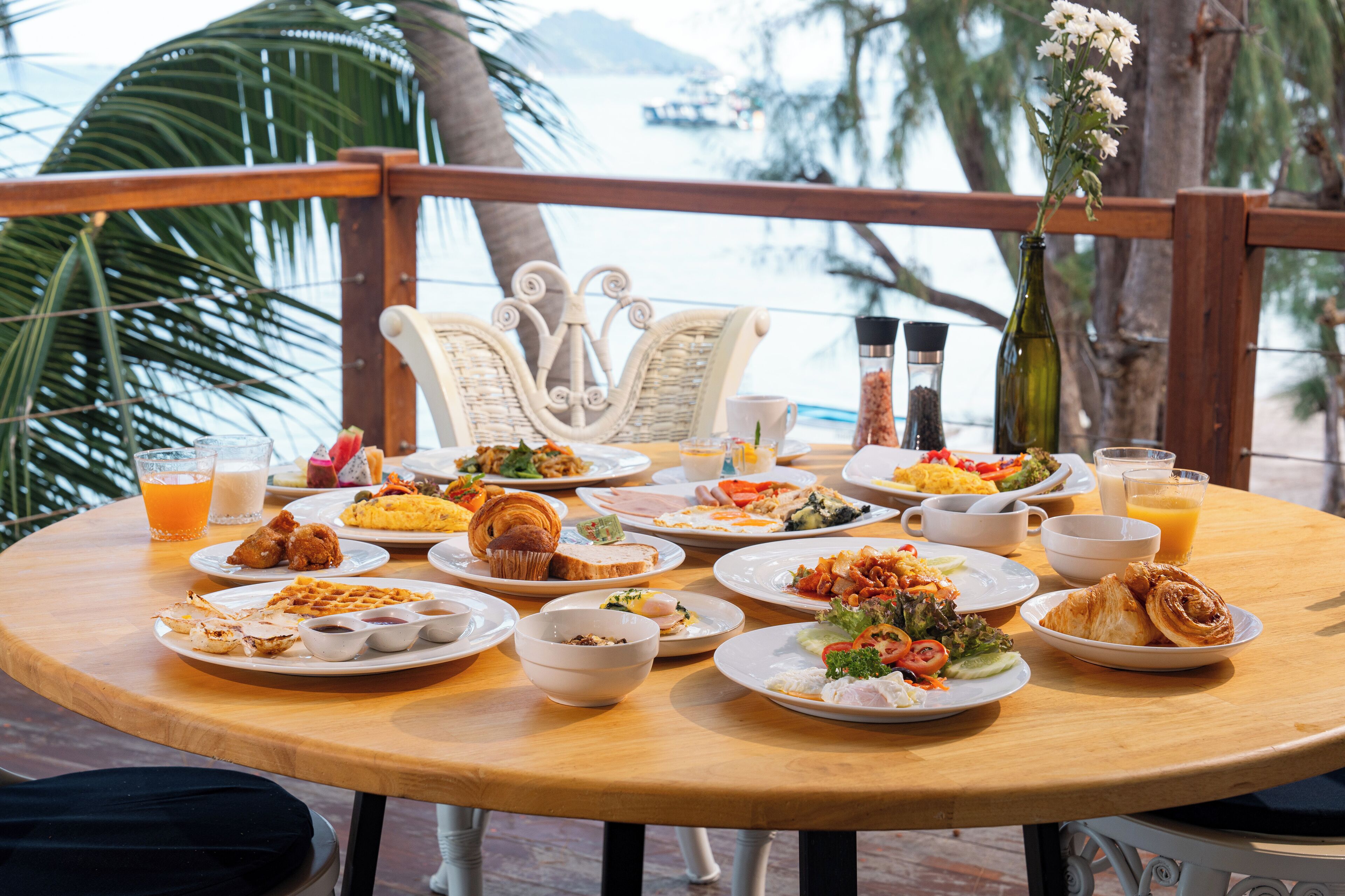Una mesa redonda de madera con varios platos de desayuno, incluyendo cruasanes, huevos y fruta, ubicada en un balcón con vistas al mar y palmeras.