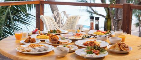 Una mesa redonda de madera con varios platos de desayuno, incluyendo cruasanes, huevos y fruta, ubicada en un balcón con vistas al mar y palmeras.