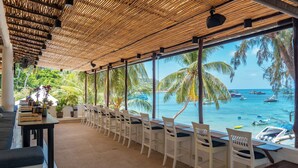 Een restaurant aan het strand met een lange tafel en stoelen, een rieten dak en uitzicht op zee en boten.