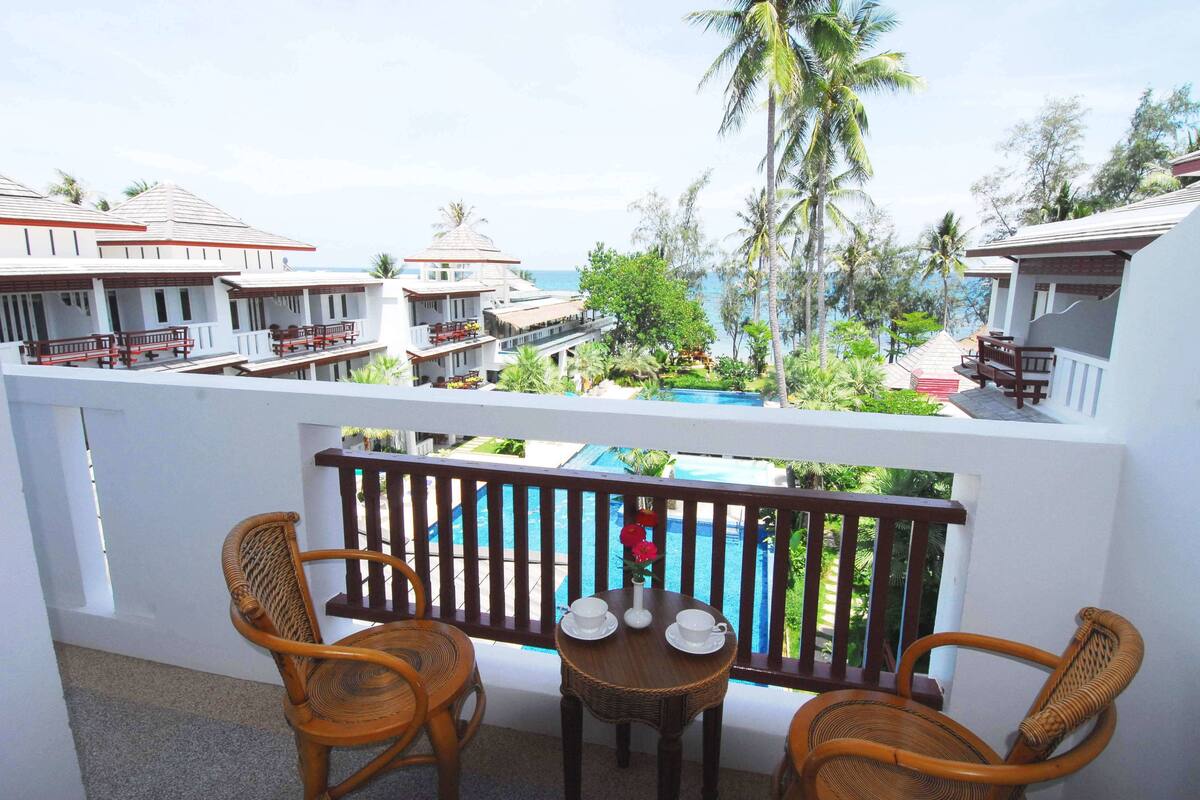 A balcony with wicker chairs and a table, overlooking a pool and resort buildings.