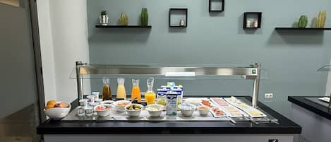 Montaje tipo bufé con diversos alimentos y bebidas, una máquina de café y un refrigerador.