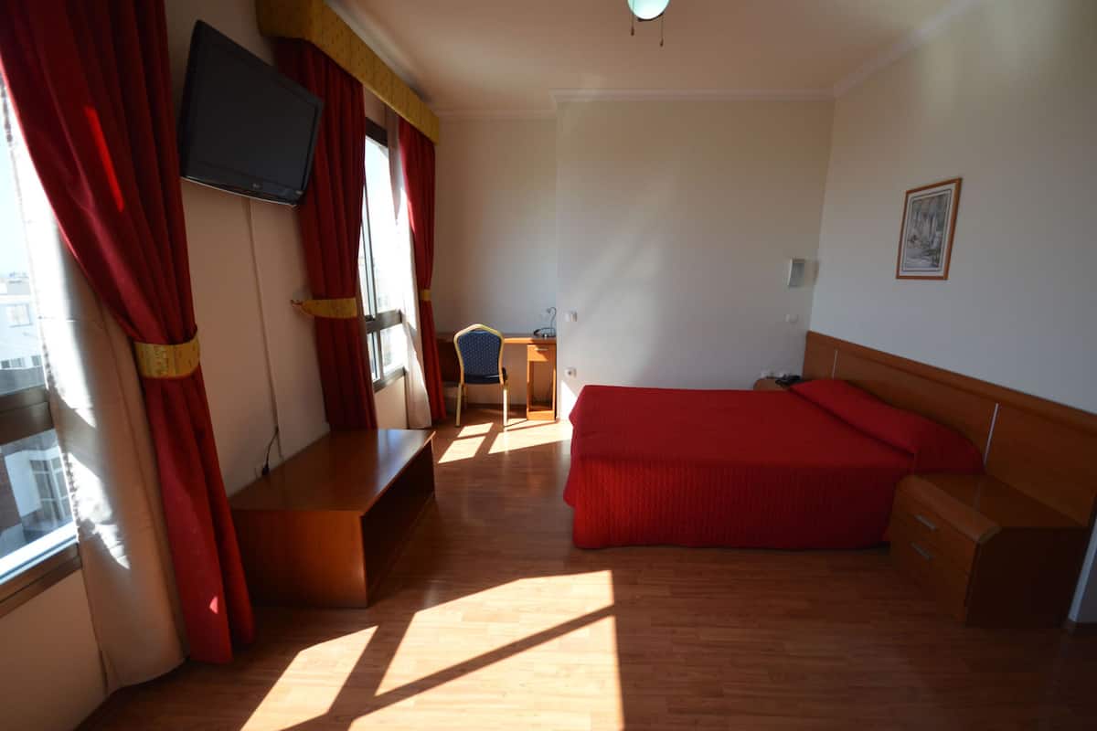 Ein Schlafzimmer mit Bett, Schreibtisch, Stuhl, Fernseher und einem Fenster mit Vorhängen.