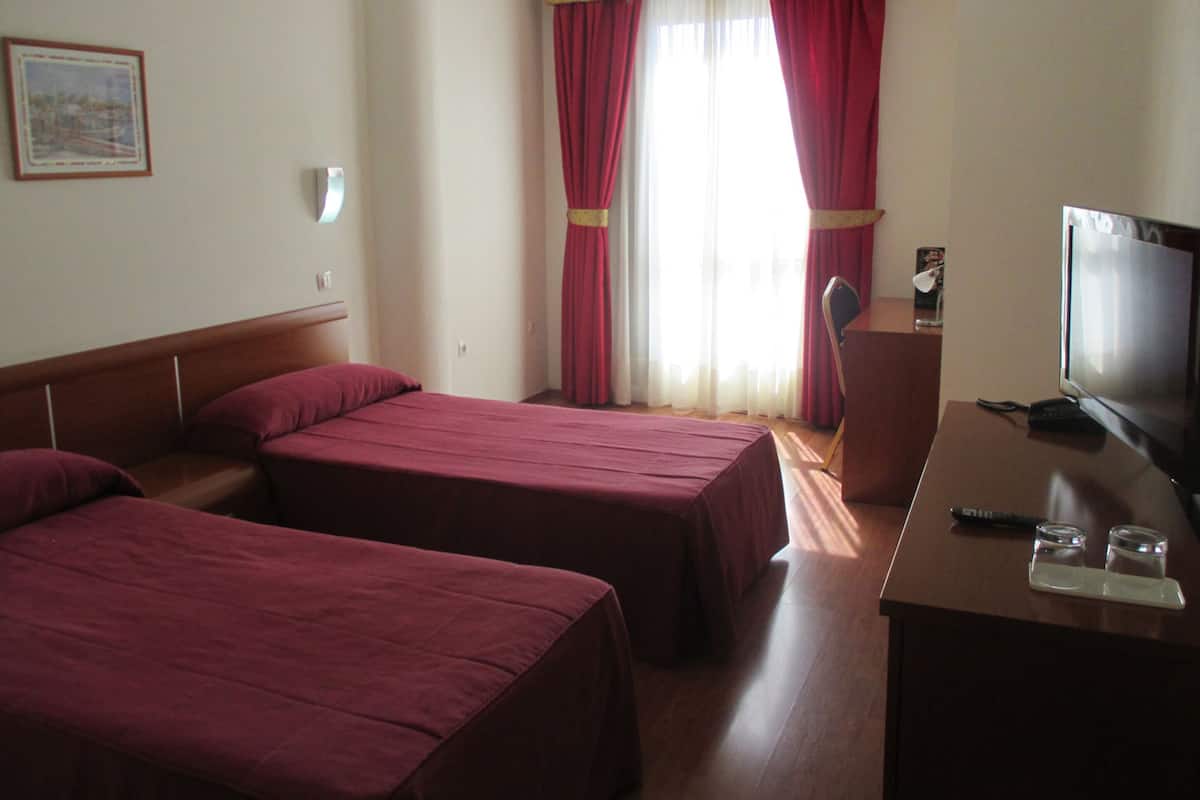 Ein Hotelzimmer mit zwei Einzelbetten, einem Schreibtisch mit Fernseher und einem Fenster mit roten Vorhängen.