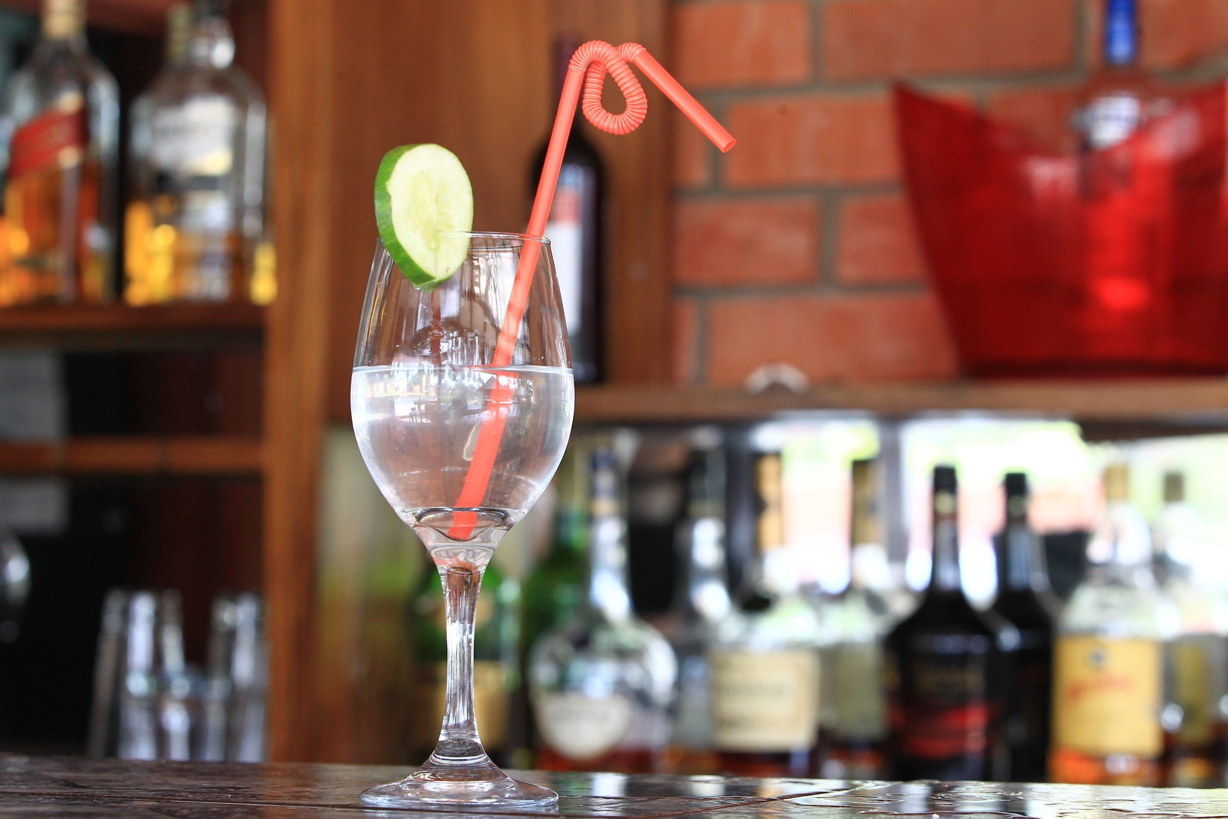 A clear glass with a lime slice and a straw, placed on a bar counter with bottles in the background.