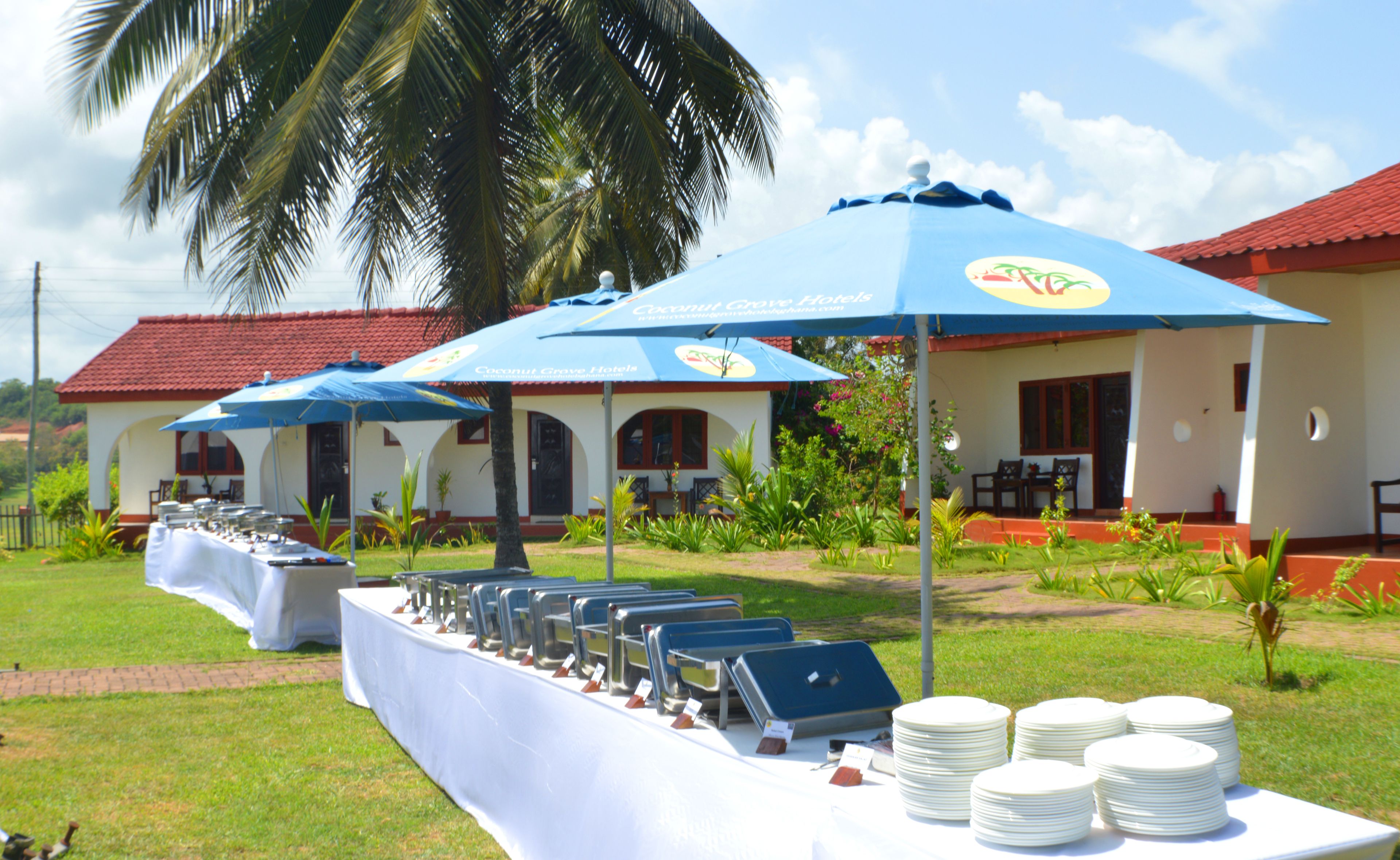 Un hôtel avec une terrasse où l’on peut déjeuner, abritée par des parasols bleus, une table blanche sur laquelle sont empilées des assiettes, et un palmier en arrière-plan.