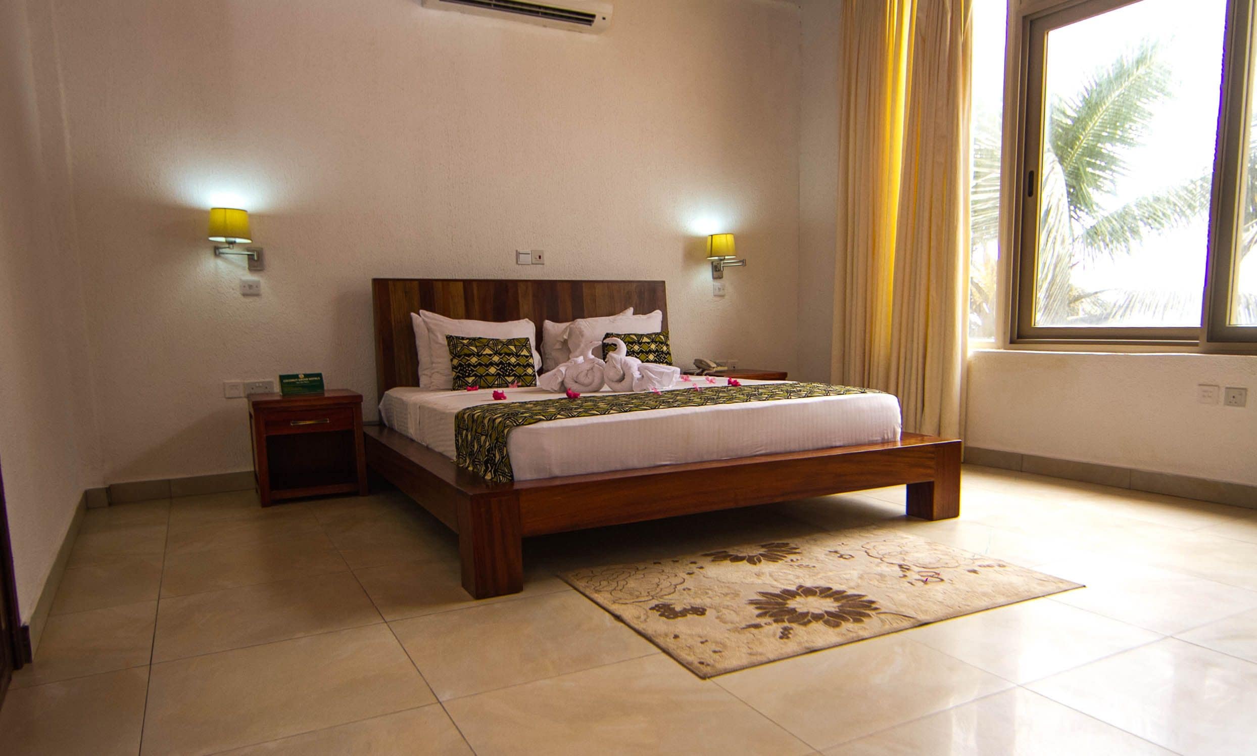 Habitación de hotel con cama de madera, dos mesitas de noche, una lámpara de noche, alfombra decorativa y ventana con cortinas.
