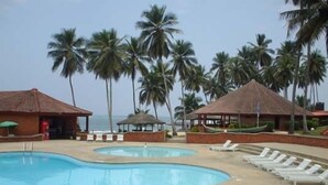 Un área de piscina con sillones de descanso, palmeras y estructuras de techo de paja.