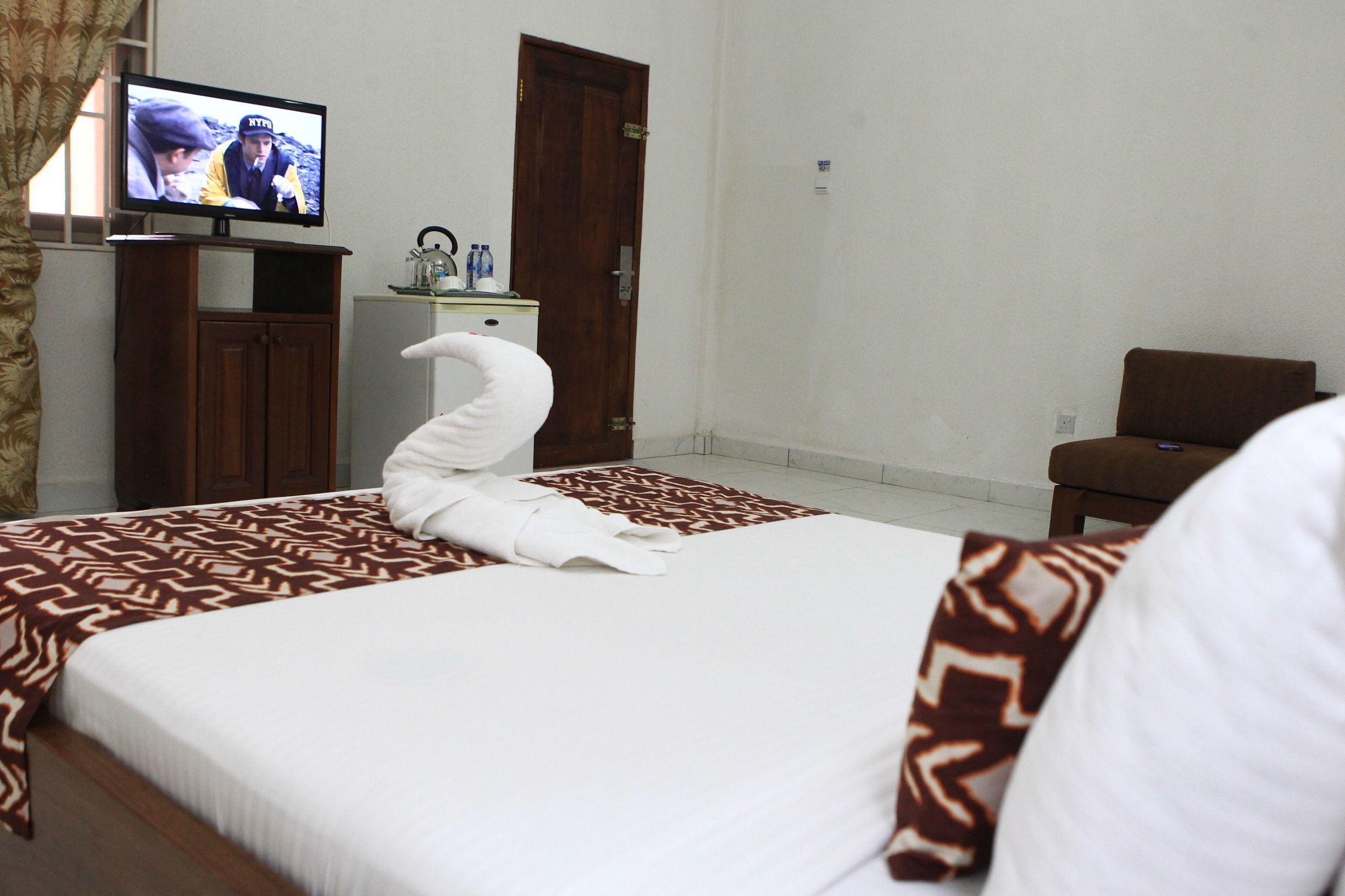 A hotel room with a neatly made bed, a television on a stand, a small table with bottles, and a brown chair.