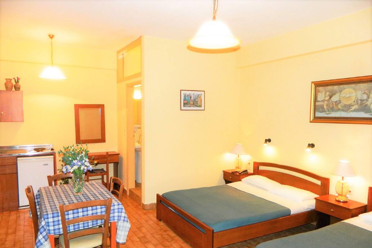 A hotel room with two single beds, a small dining table with chairs, a kitchenette, and a painting on the wall.