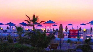 Resort à beira-mar com guarda-sóis e espreguiçadeiras, palmeiras e vista para o pôr do sol.