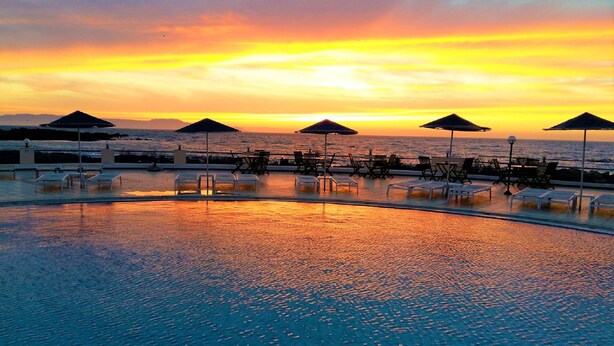 Área da piscina com espreguiçadeiras e guarda-sóis, com vista para o oceano ao pôr do sol.