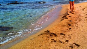 Uma mulher em uma praia arenosa, com os braços levantados, olhando para as montanhas e o mar.