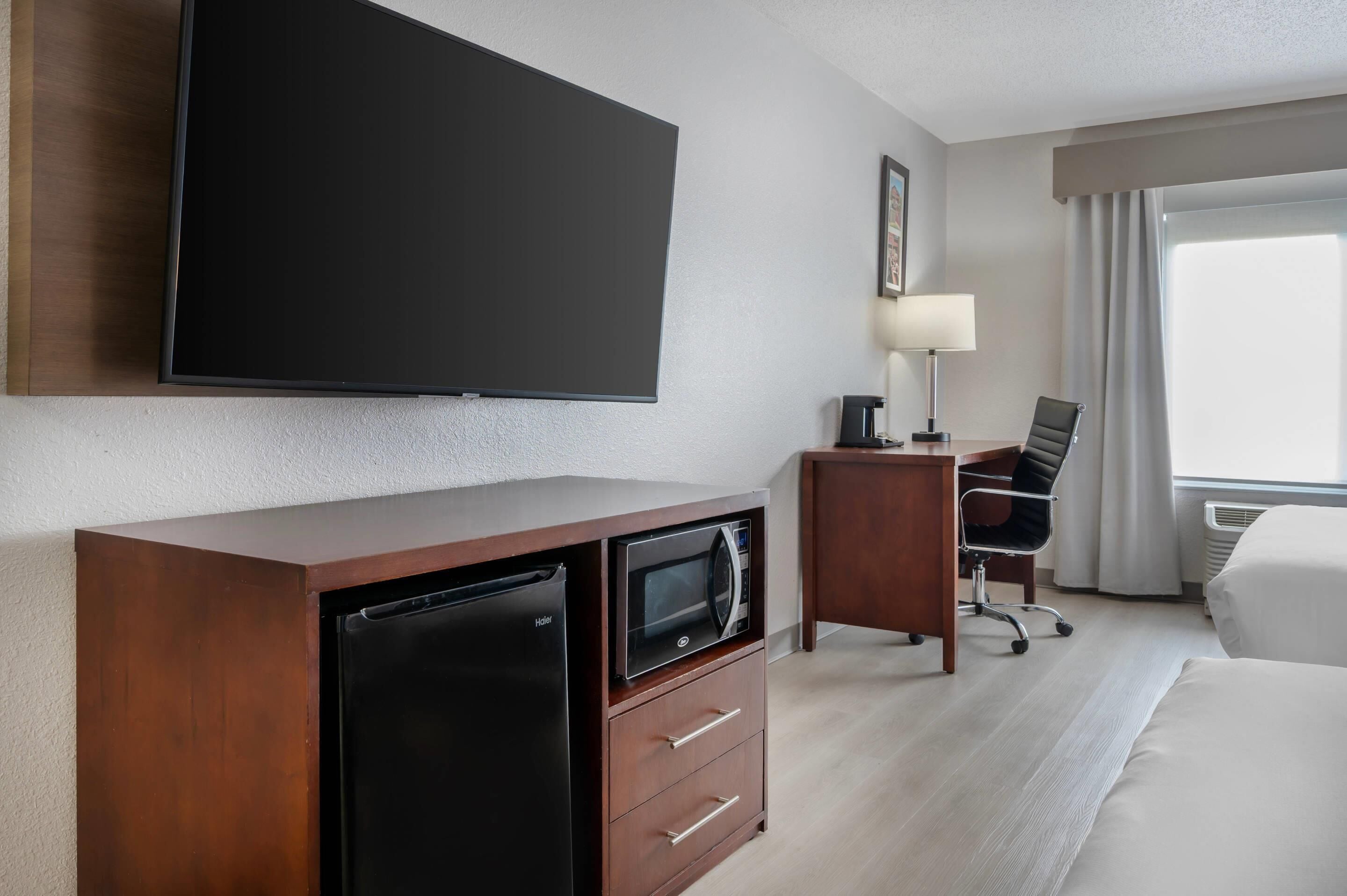 A hotel room with a flat-screen TV, a minibar, a desk with a lamp, a chair, and a bed.
