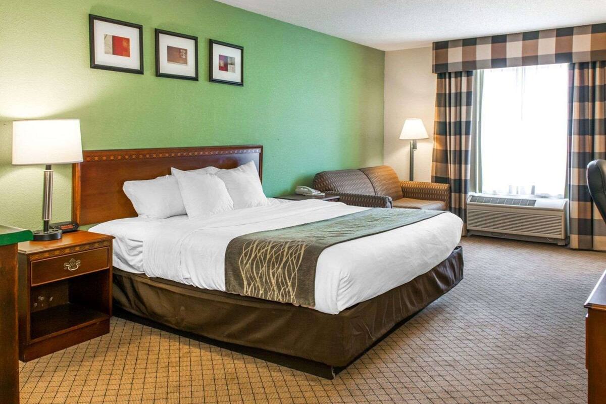 A hotel room with a large bed, a bedside table with a lamp, a chair, a desk, and a window with curtains.