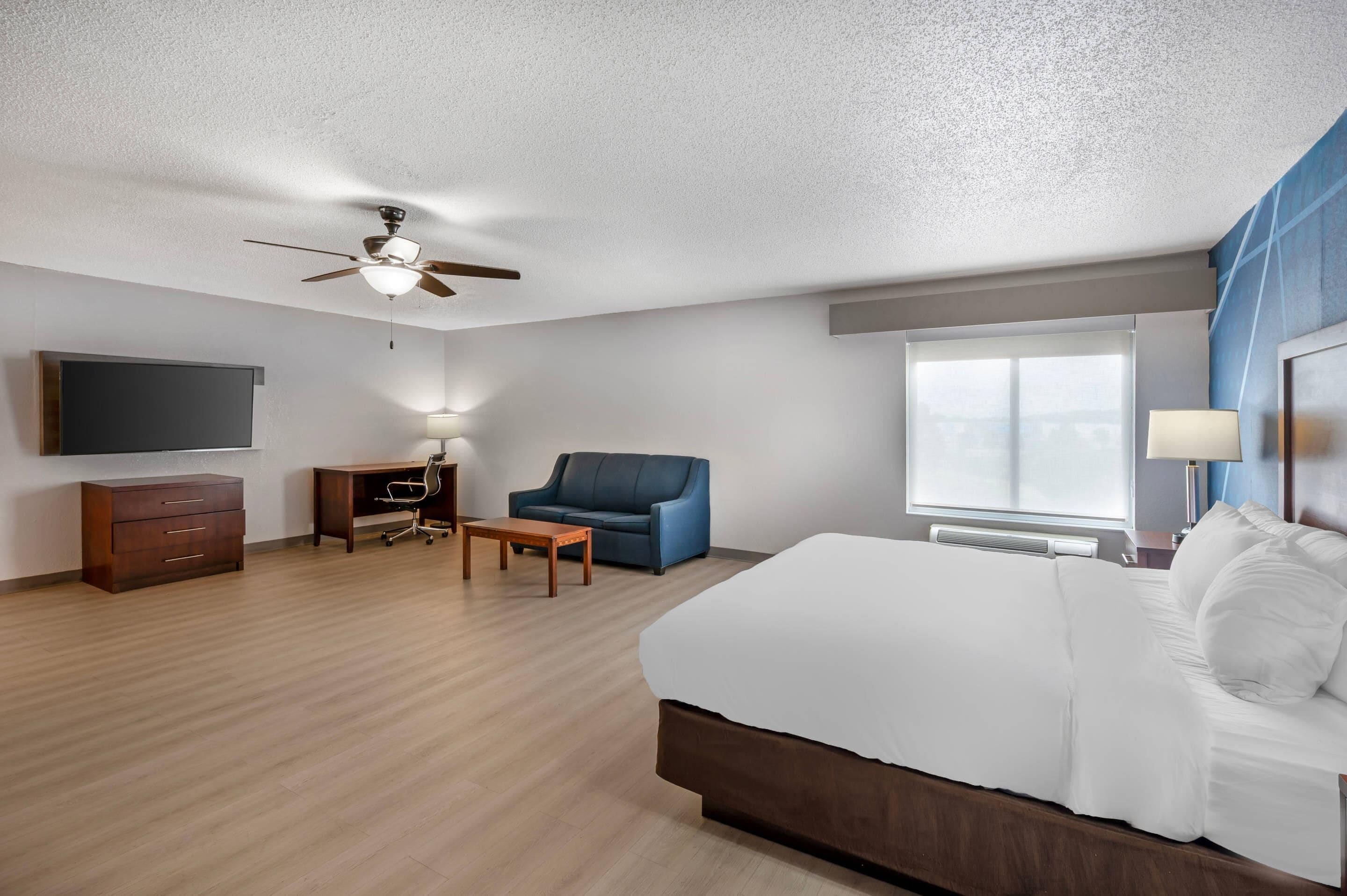 A hotel room with a bed, a desk, a chair, a ceiling fan, a TV, and a window.