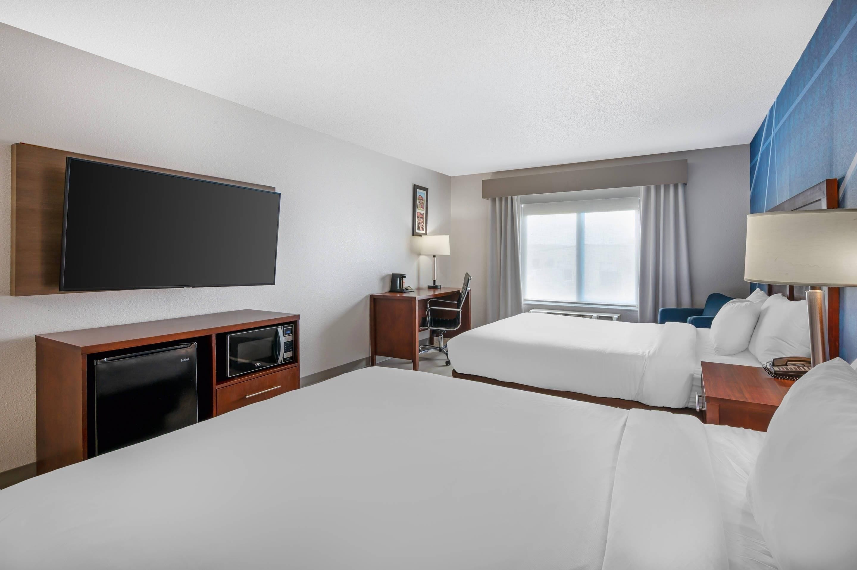 Quarto de hotel com duas camas, televisão de tela plana, escrivaninha com computador, abajur e janela com cortinas.