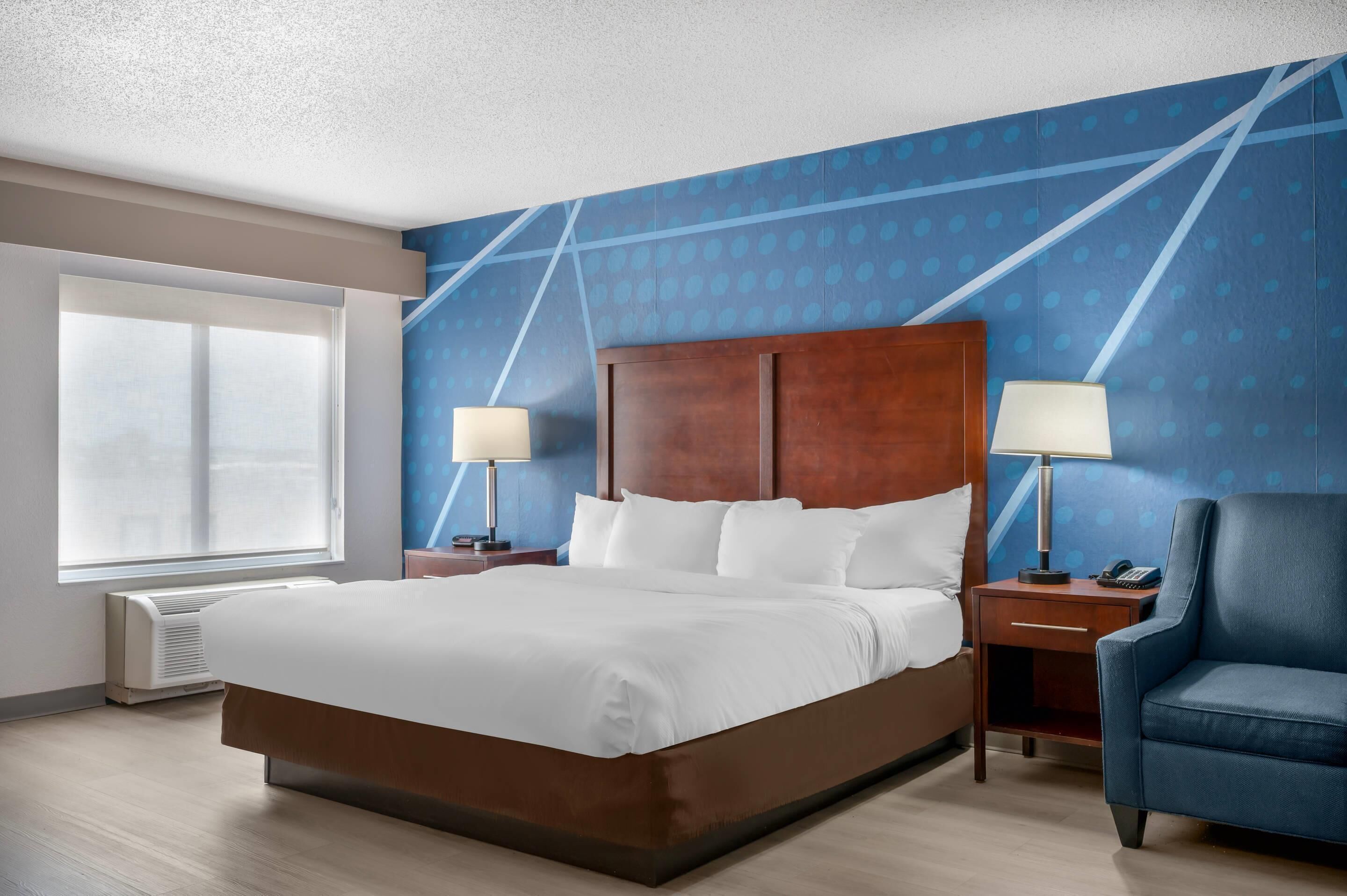 A hotel room with a large bed, two bedside tables with lamps, a chair, a large window, and a blue patterned wall.