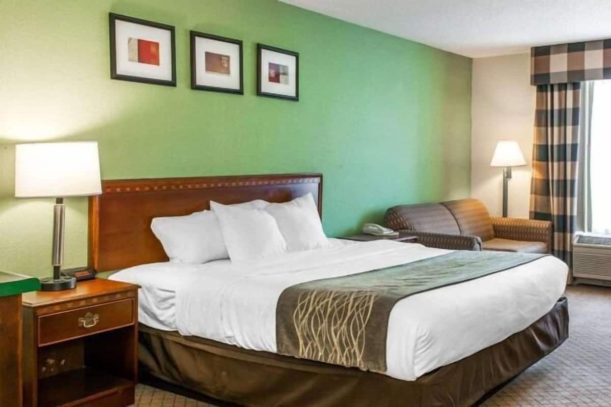 A hotel room with a bed, bedside table, lamp, chair, and air conditioning unit.