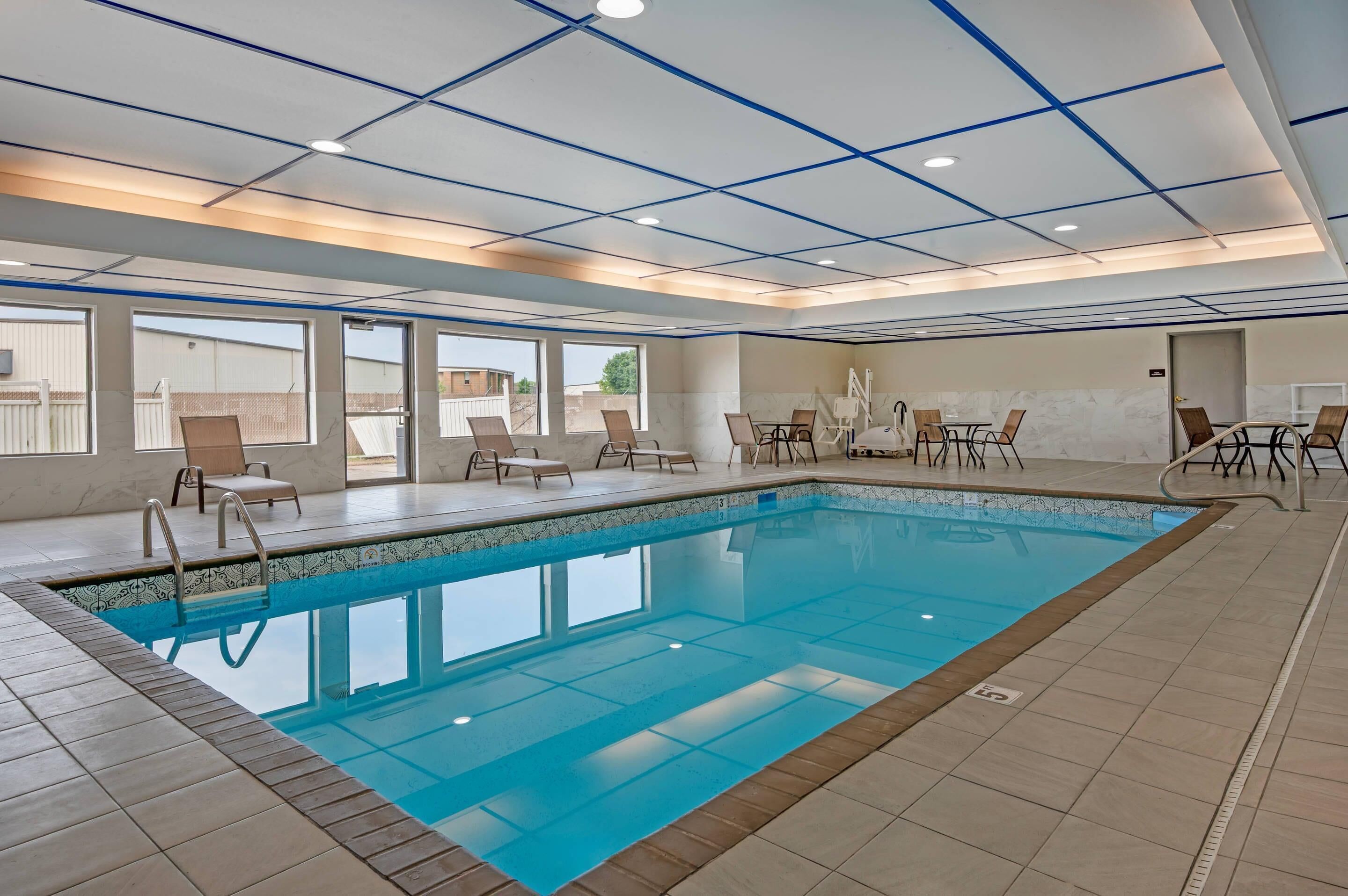 A swimming pool with lounge chairs and tables, surrounded by large windows and a tiled floor.