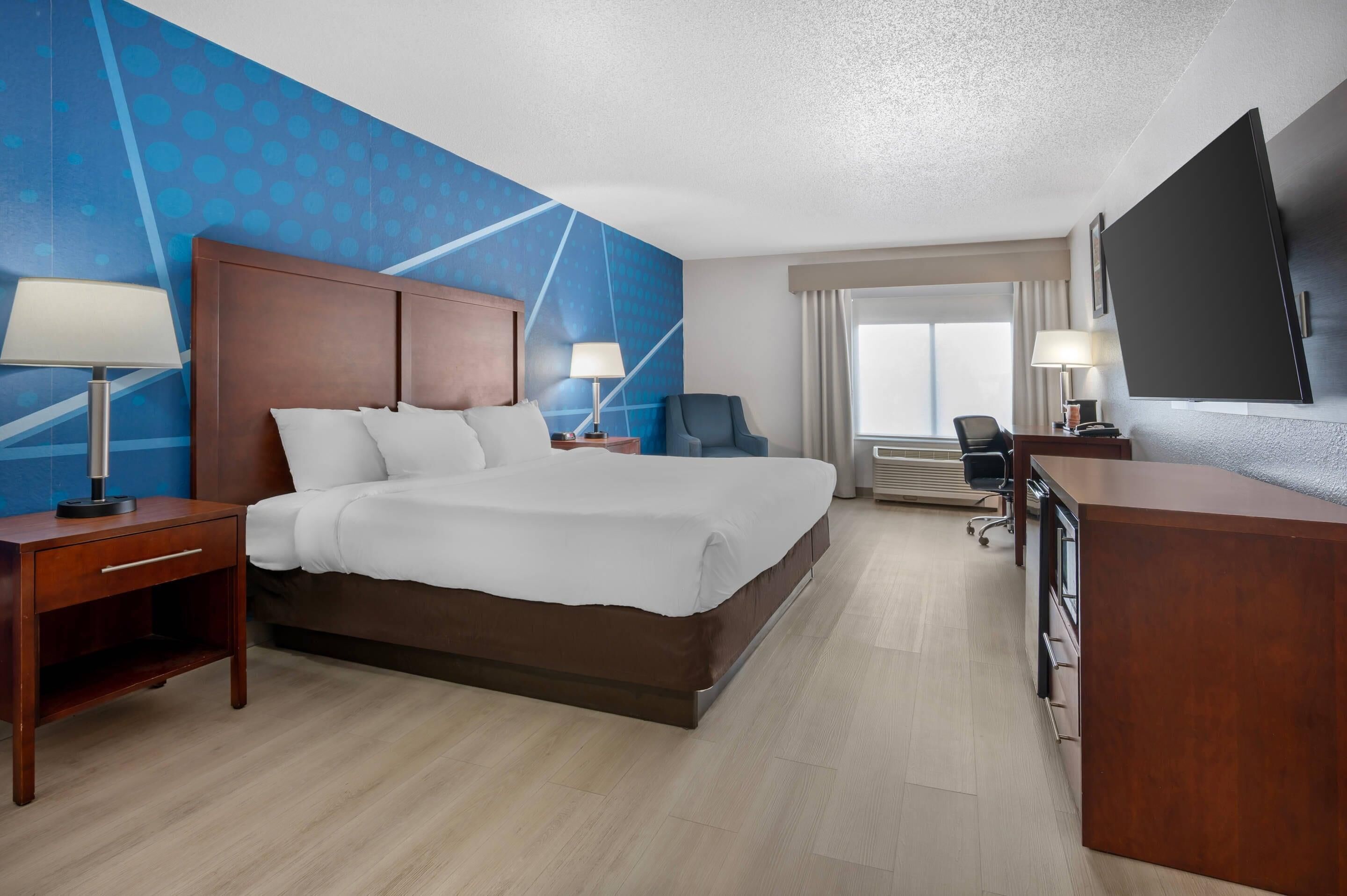A hotel room with a large bed, two bedside tables with lamps, a desk with a chair, a flat-screen TV mounted on the wall, and a window with curtains.