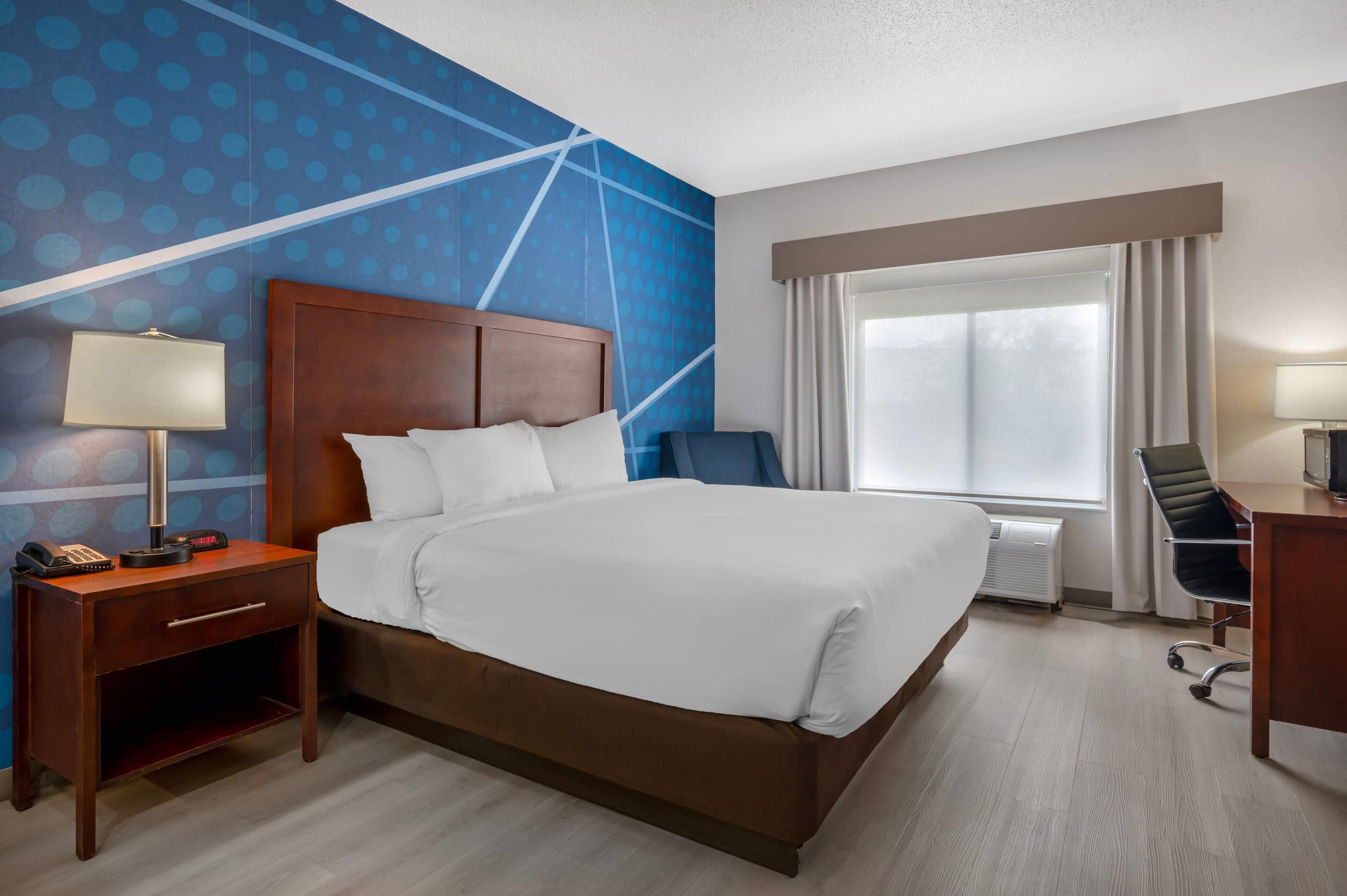 A hotel room with a bed, a desk, a chair, a lamp, and a window with curtains.