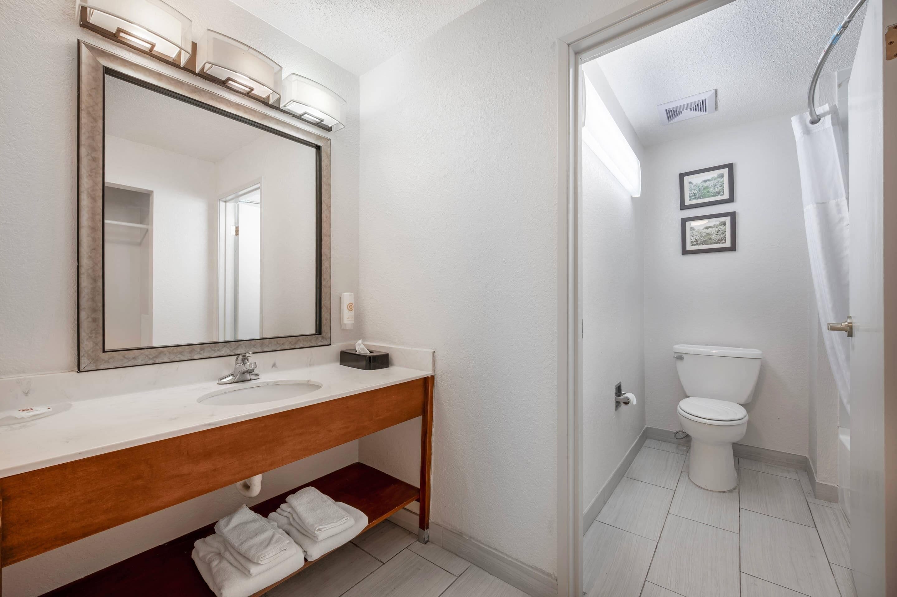 A bathroom with a white sink, mirror, and toilet. There is a towel rack with folded towels and a small shelf below the sink.