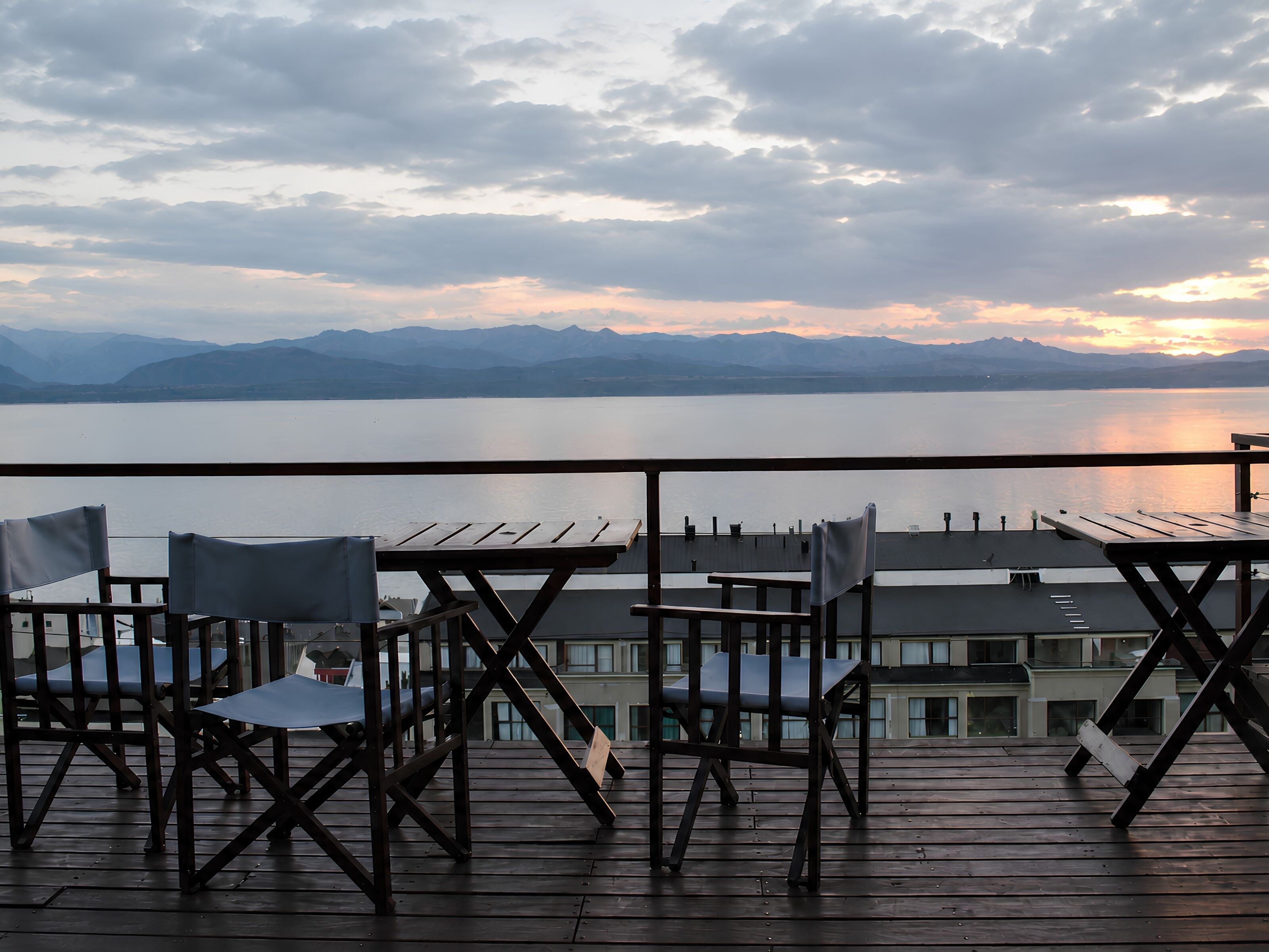 A wooden deck with chairs and tables overlooking a calm lake with mountains in the distance.