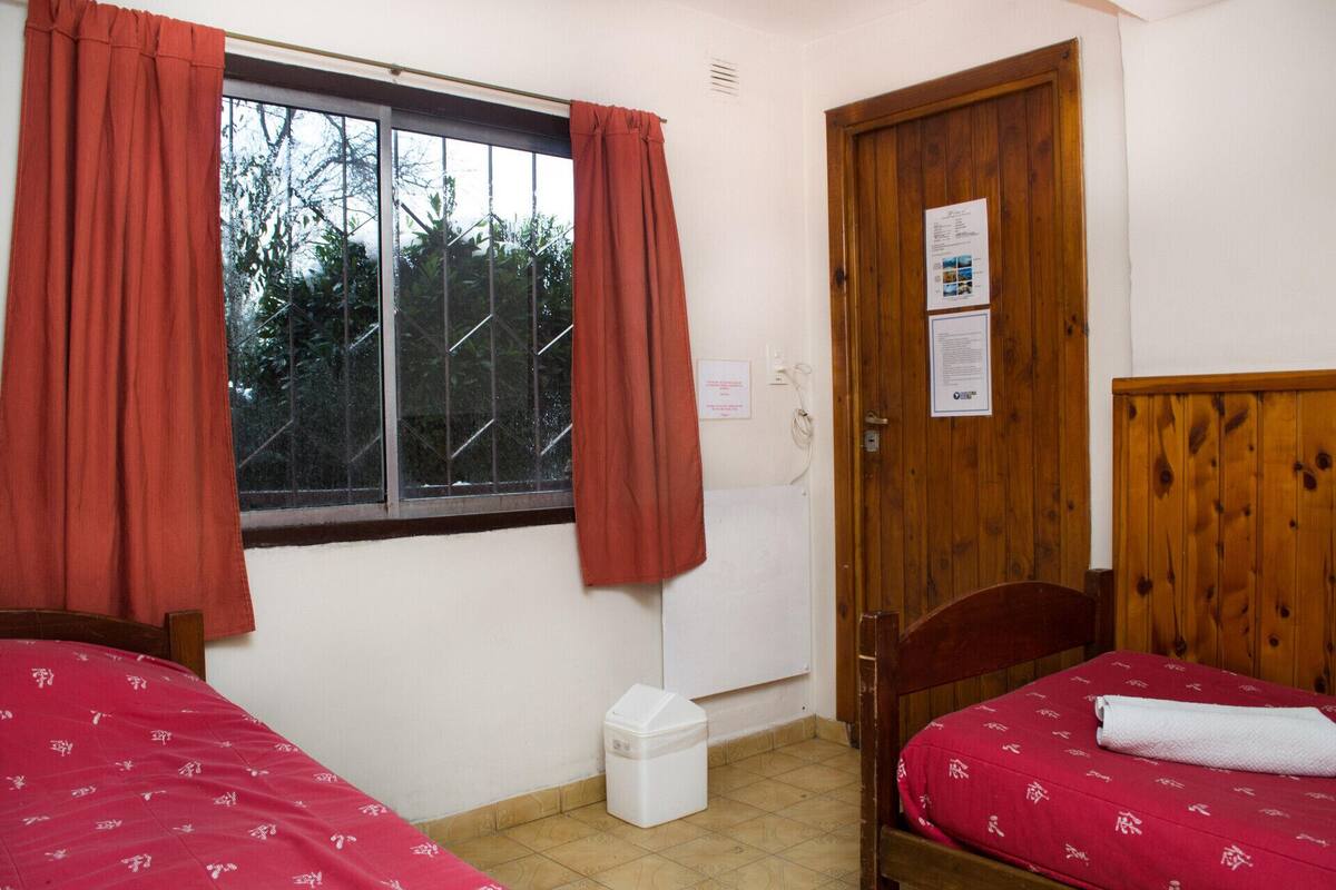 A small room with two beds, a window with curtains, a wooden door, and a white trash bin.