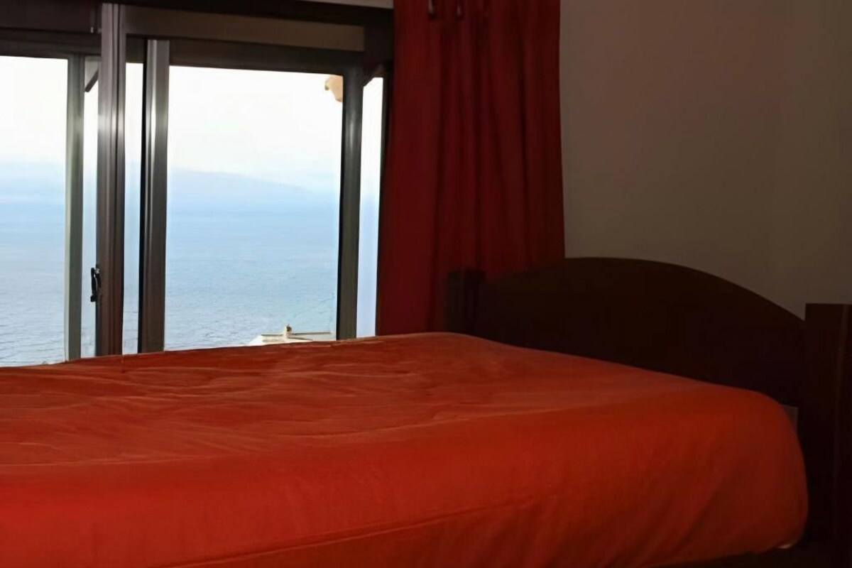 A room with a bed, a view of the sea, and a window with curtains.