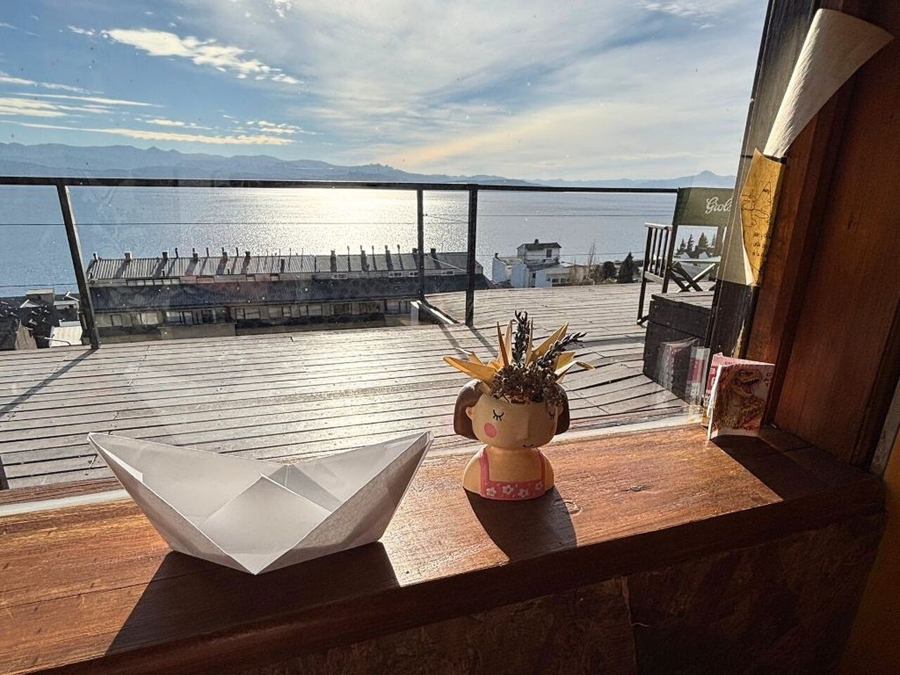 Un balcón con vista al mar, una mesa de madera con un barquito de papel y una maceta de cerámica con plantas, además de una silla.