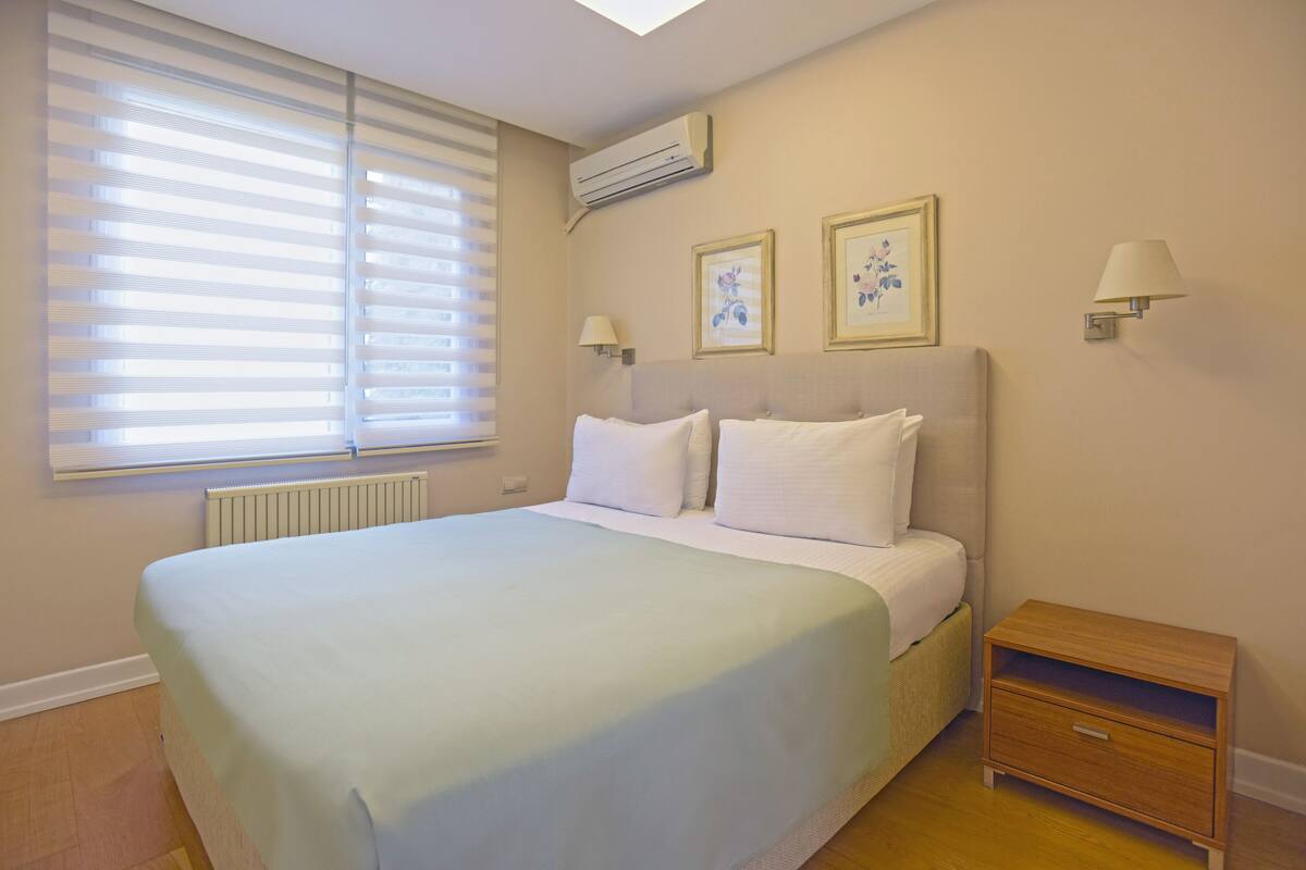 Ein ordentlich bezogenes Bett mit weißen Laken, ein Nachttisch mit Lampe sowie zwei gerahmte Bilder an der Wand.
