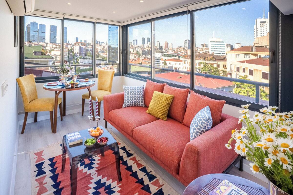 Ein modernes Wohnzimmer mit einer Couch, Sesseln, einem Couchtisch und Blick auf eine Stadtlandschaft durch große Fenster.