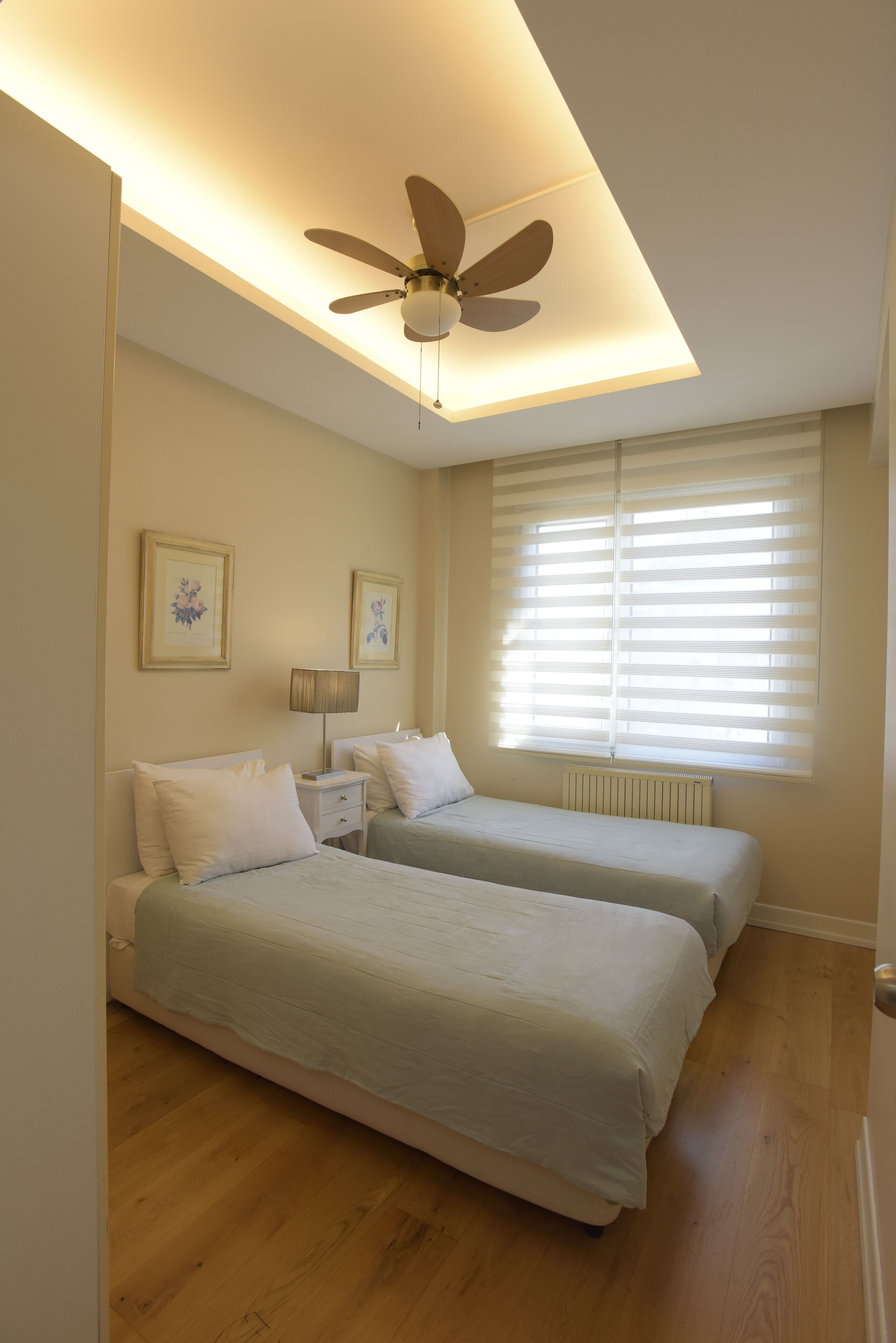 A bedroom with two beds, a ceiling fan, a window with blinds, and a radiator.
