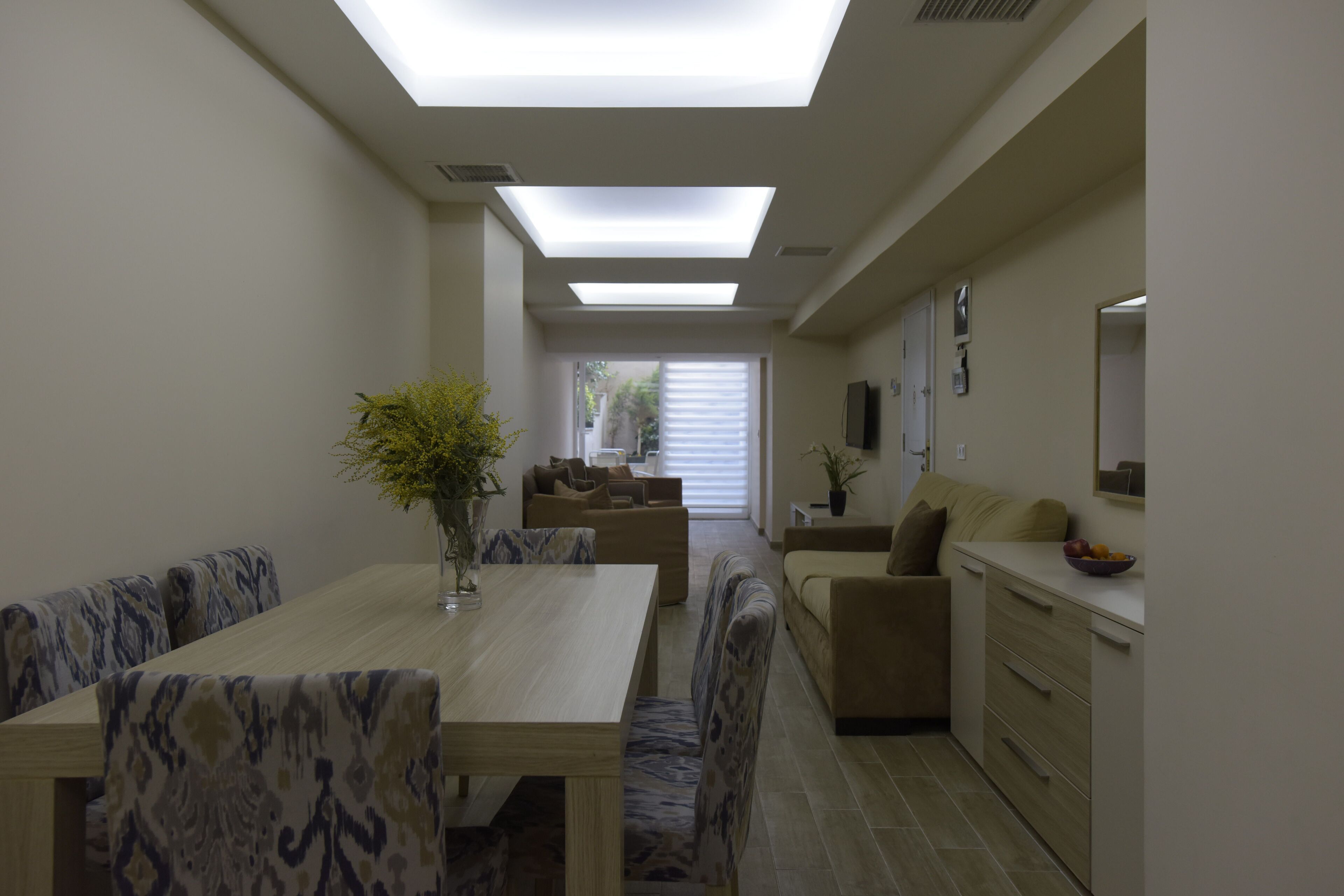 Un comedor moderno con una mesa larga y sillas estampadas, un sofá, y una zona de cocina con mostrador y fregadero.