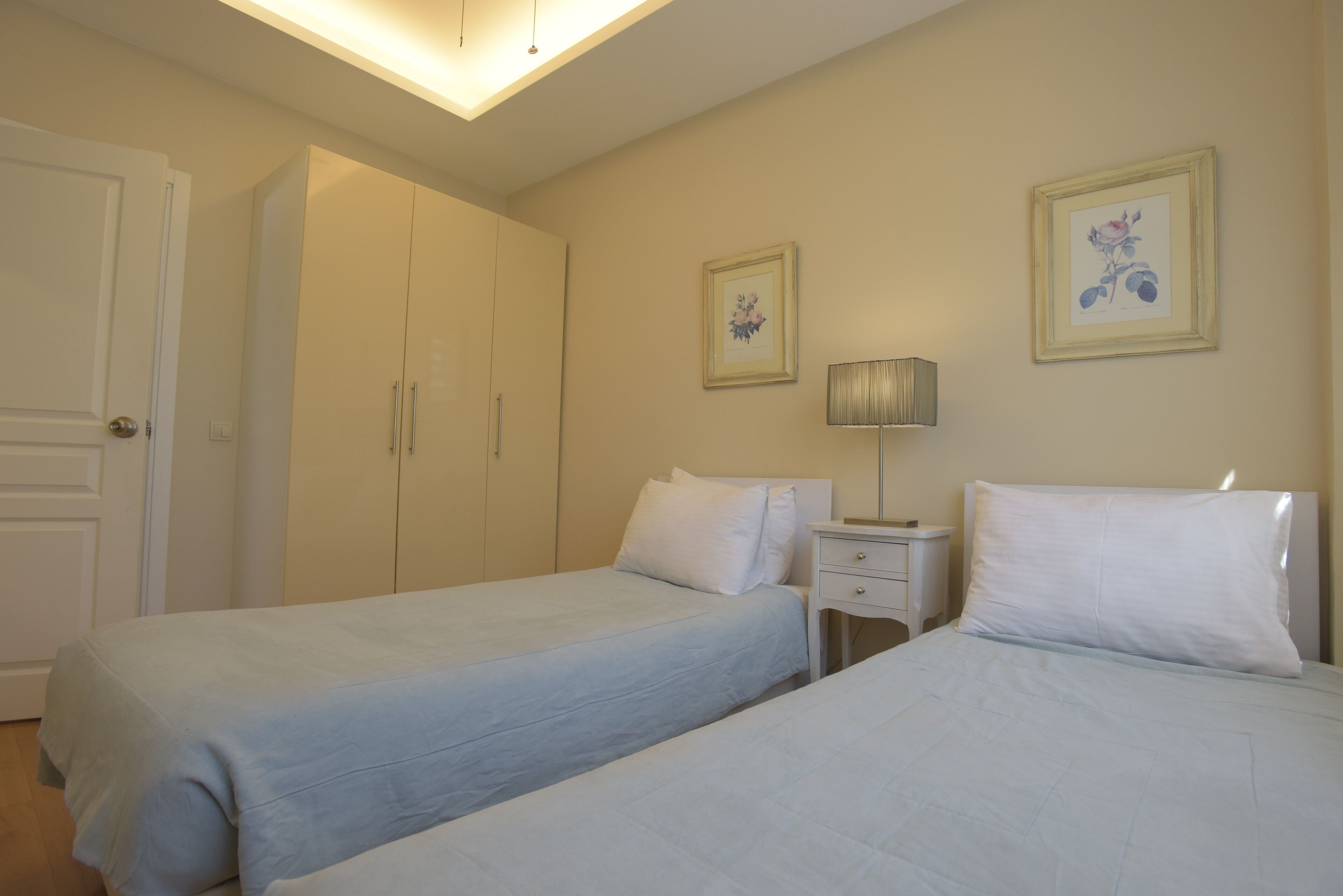A bedroom with two beds, a wardrobe, a bedside table with a lamp, and two framed pictures on the wall.