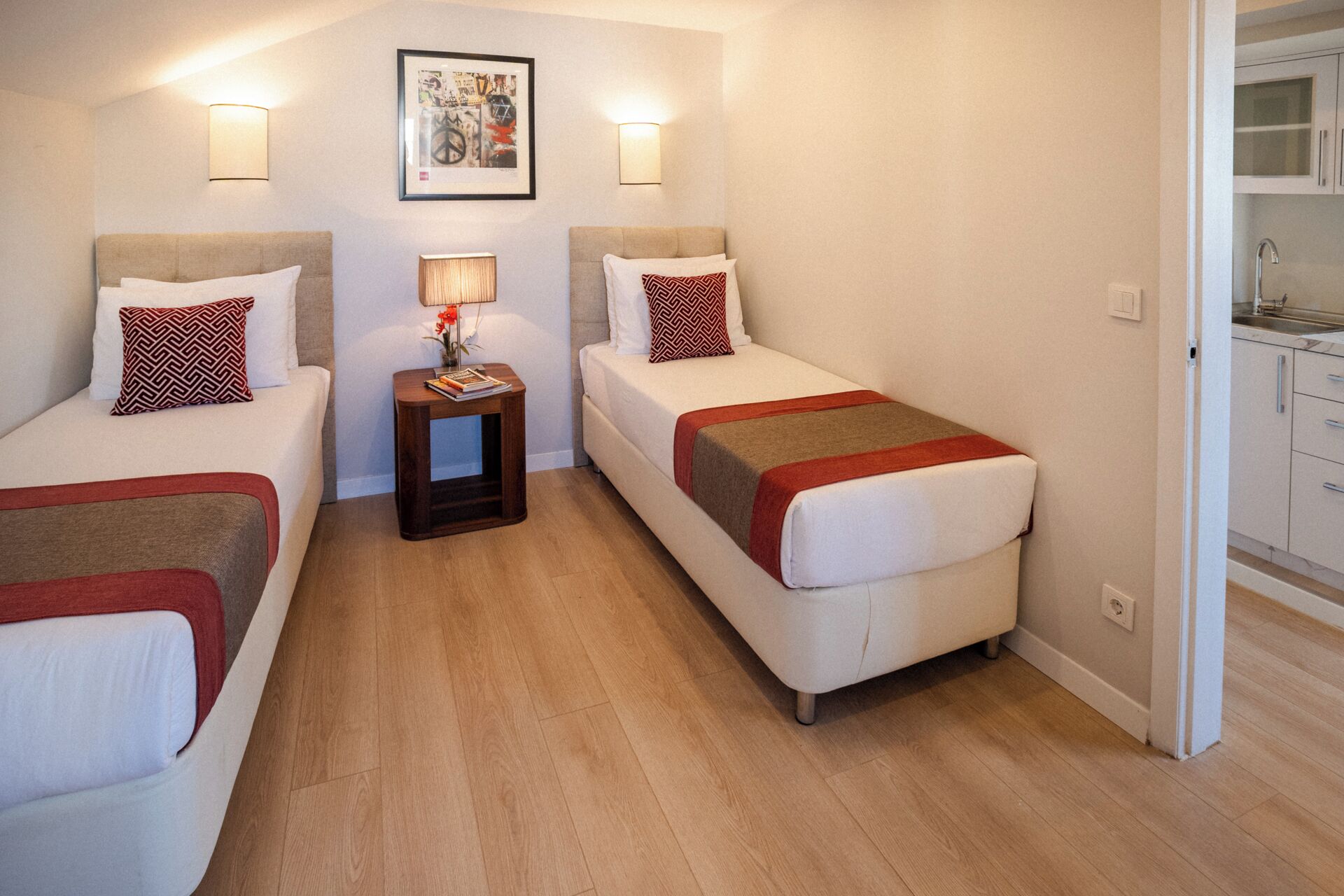 Quarto de hotel pequeno e moderno, com duas camas de solteiro, mesa de cabeceira, abajur e um quadro na parede.
