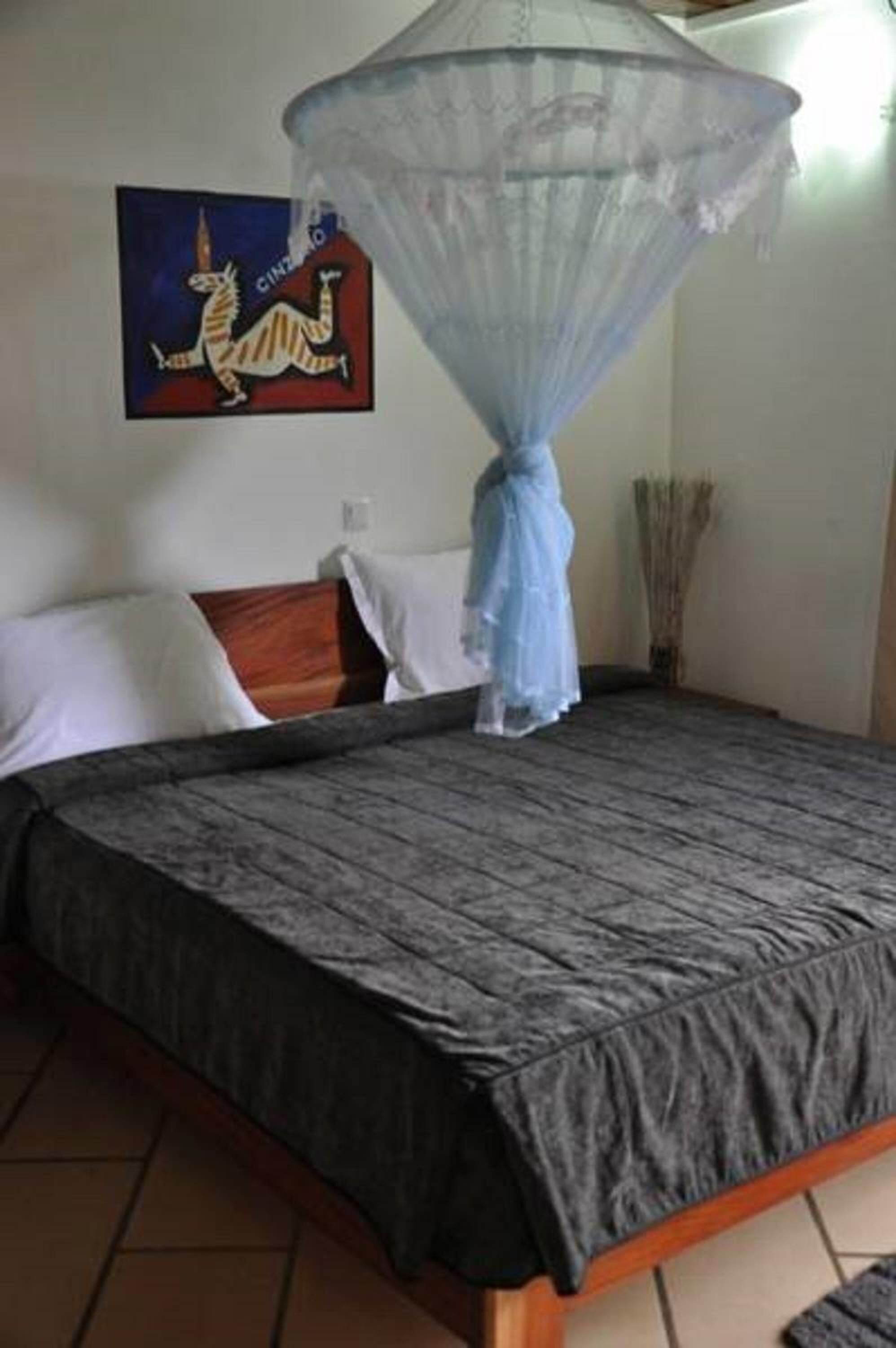 A bedroom with a bed, a mosquito net, a painting, and a vase with reeds.