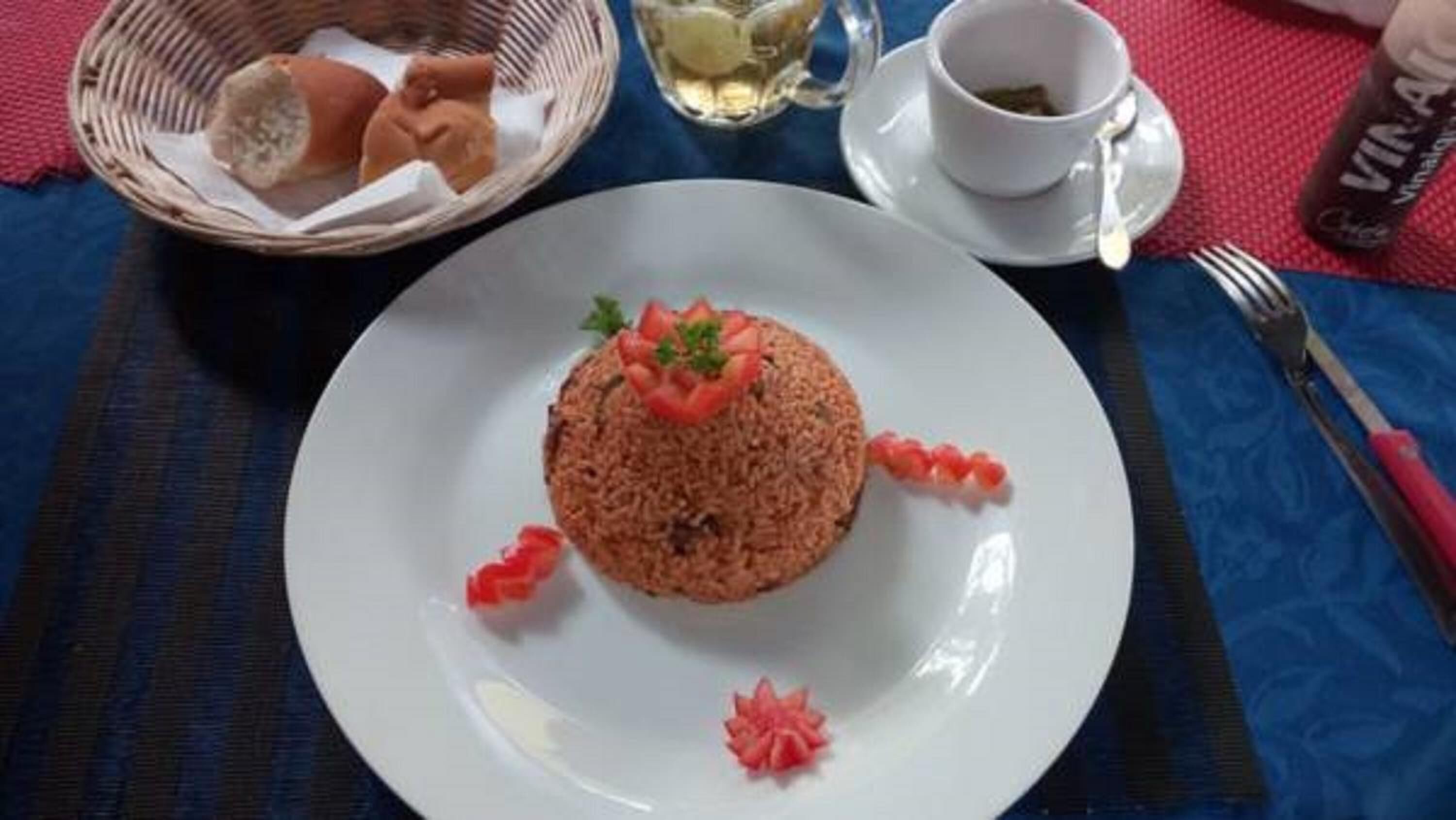Un piatto di cibo con una composizione decorativa di elementi rossi e bianchi, una tazza di tè e una bottiglia d'acqua su un tavolo con tovaglia blu.