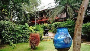 A tropical garden with a large blue vase, a small red bush, and a wooden structure in the background. - Napoleon Lagune (Lomé)