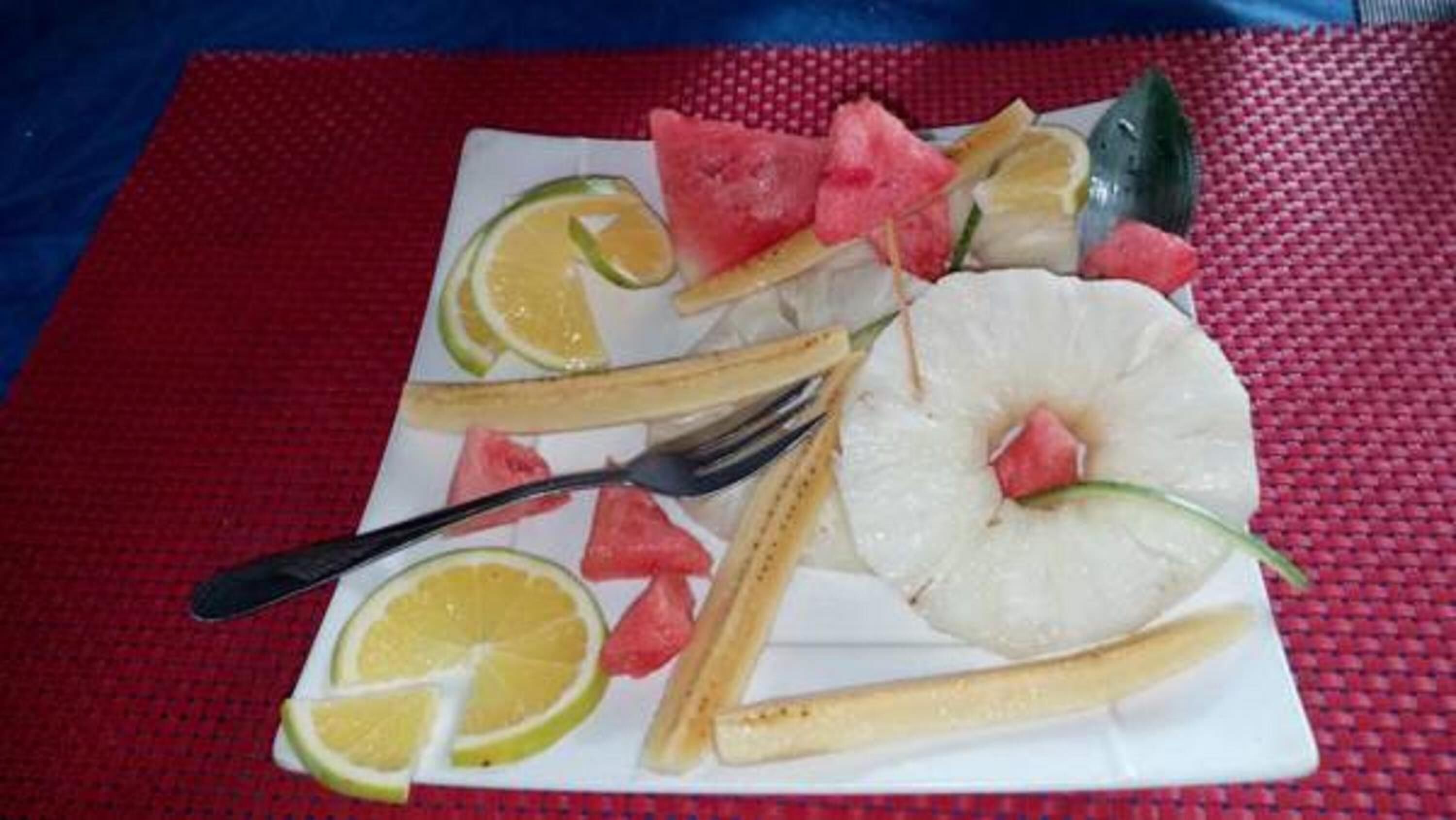 Une assiette de fruits comprenant des tranches de pastèque, d’ananas et d’orange, posée sur une nappe rouge à texture.