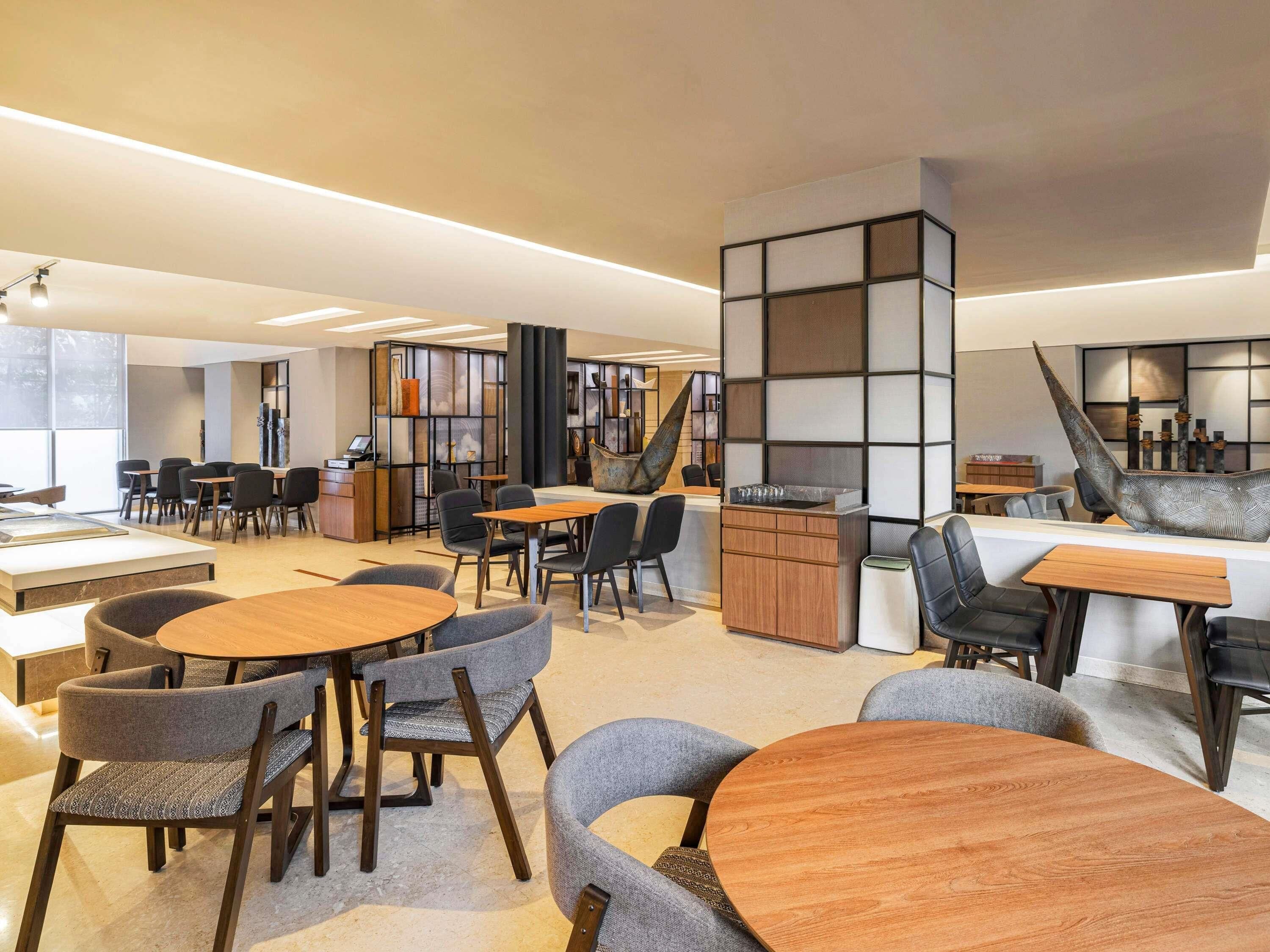 A modern interior with round wooden tables and grey upholstered chairs, a reception desk, and a decorative wall with a unique sculpture.