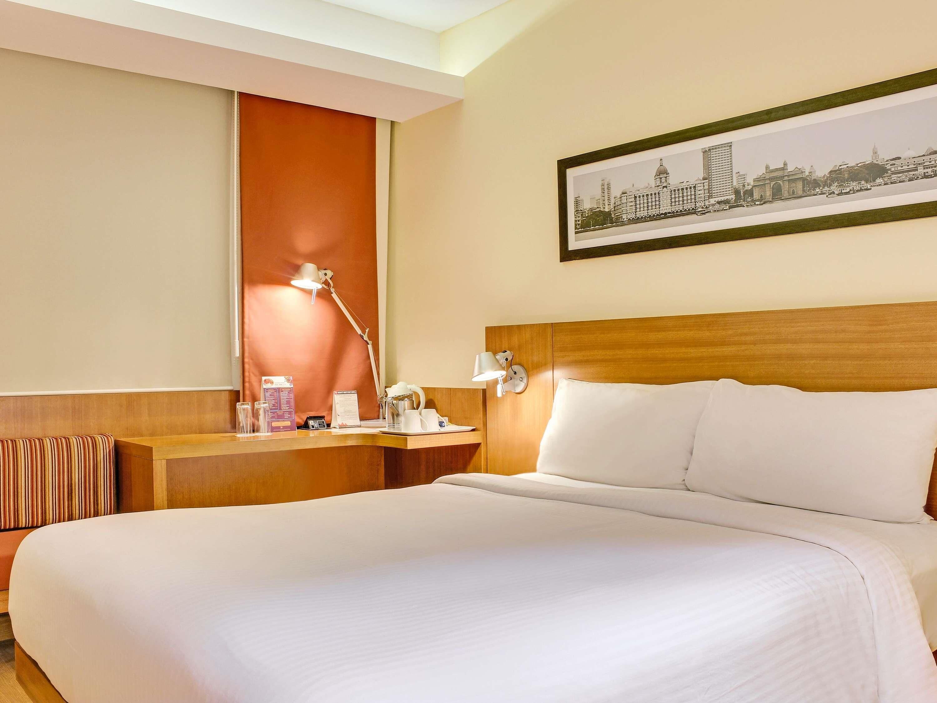 A hotel room with a large bed, a wooden headboard, a desk with a lamp, a framed cityscape picture, and a window with a view of buildings.