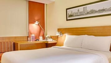 A hotel room with a large bed, a wooden headboard, a desk with a lamp, a framed cityscape picture, and a window with a view of buildings.