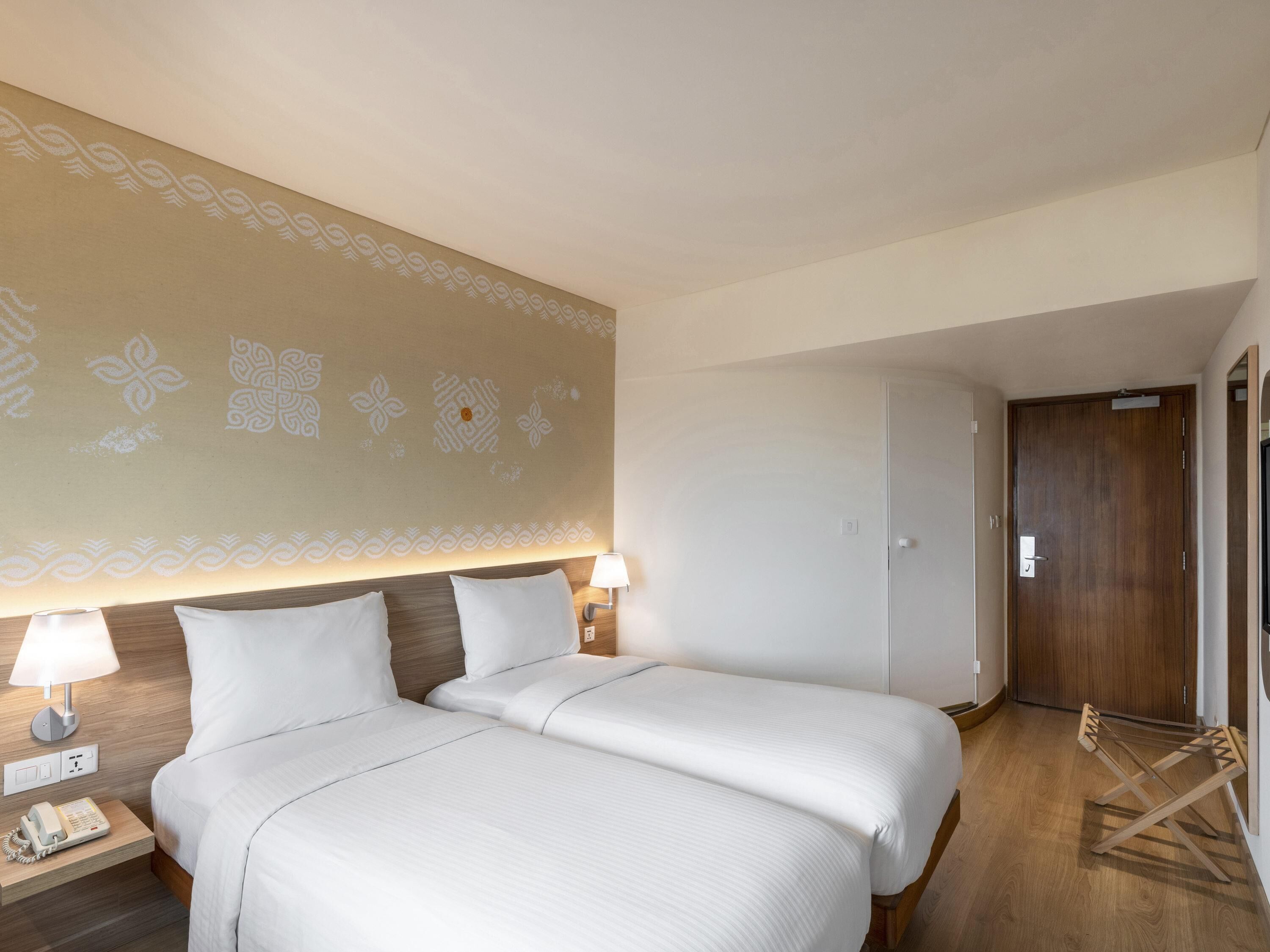 A hotel room with two beds, a wooden headboard, a wall with decorative patterns, a bedside lamp, a phone on the nightstand, and a wooden door.