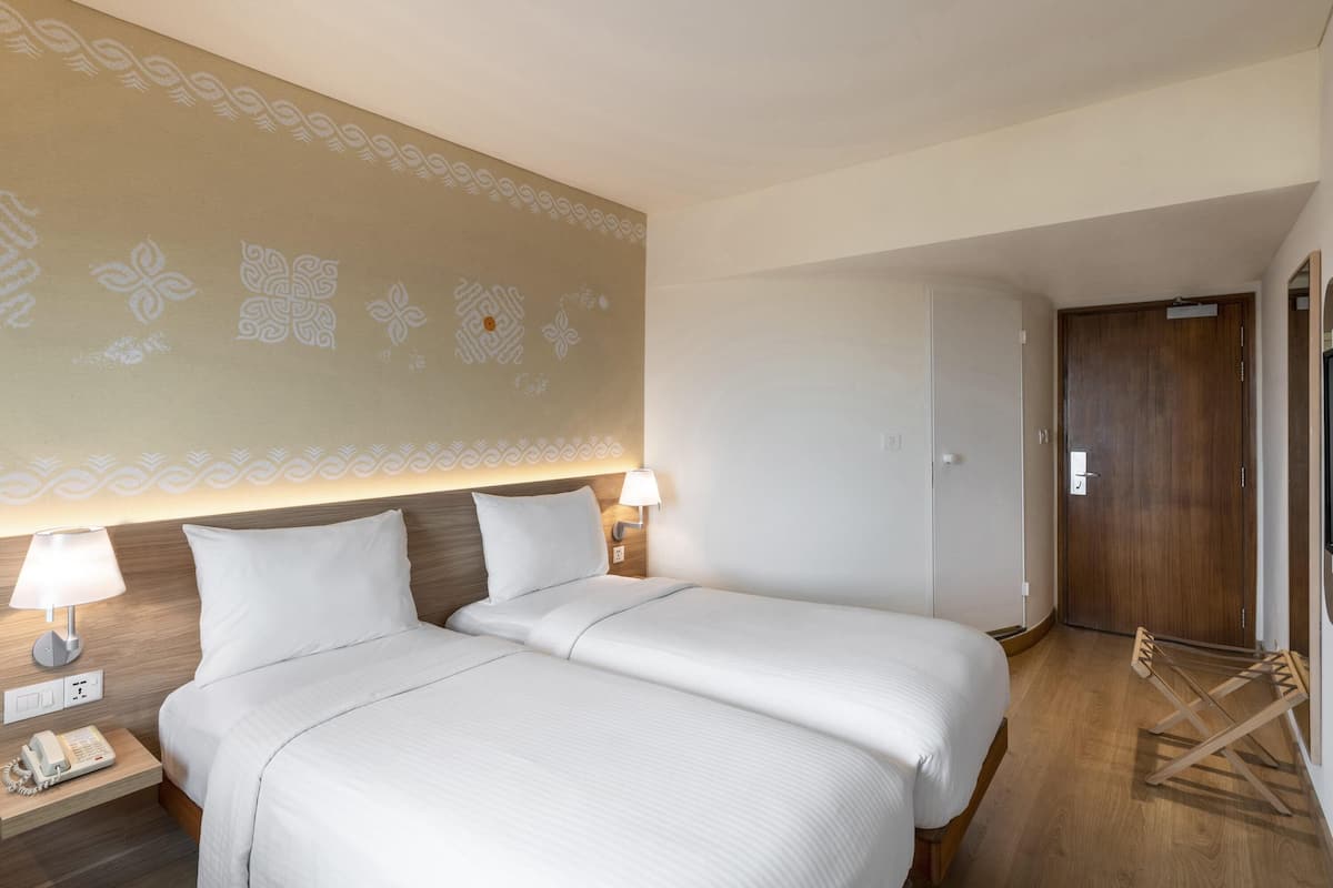 A hotel room with two beds, a wooden headboard, a wall with decorative patterns, a bedside lamp, a phone on the nightstand, and a wooden door.