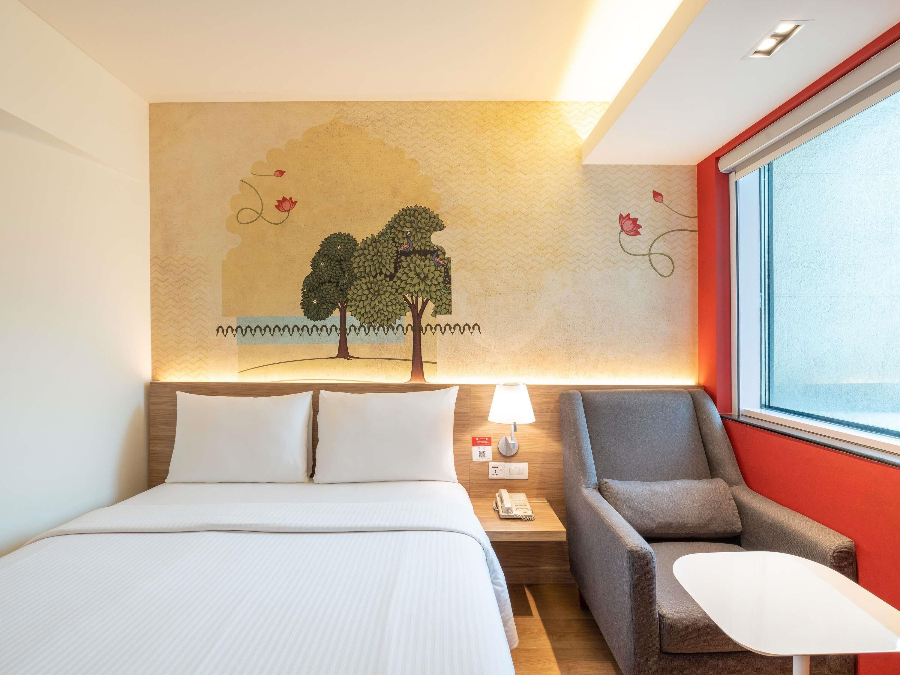 A hotel room with a bed, a desk, a chair, a lamp, and a wall mural.