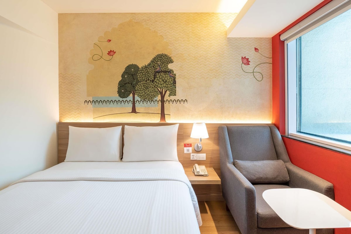 A hotel room with a bed, a desk, a chair, a lamp, and a wall mural.