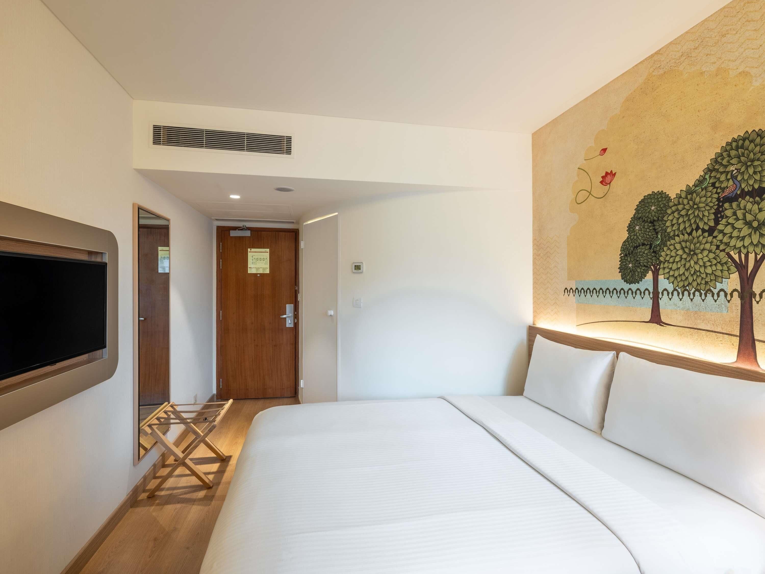 A hotel room with a bed, a TV mounted on the wall, a door, and a painting of trees.