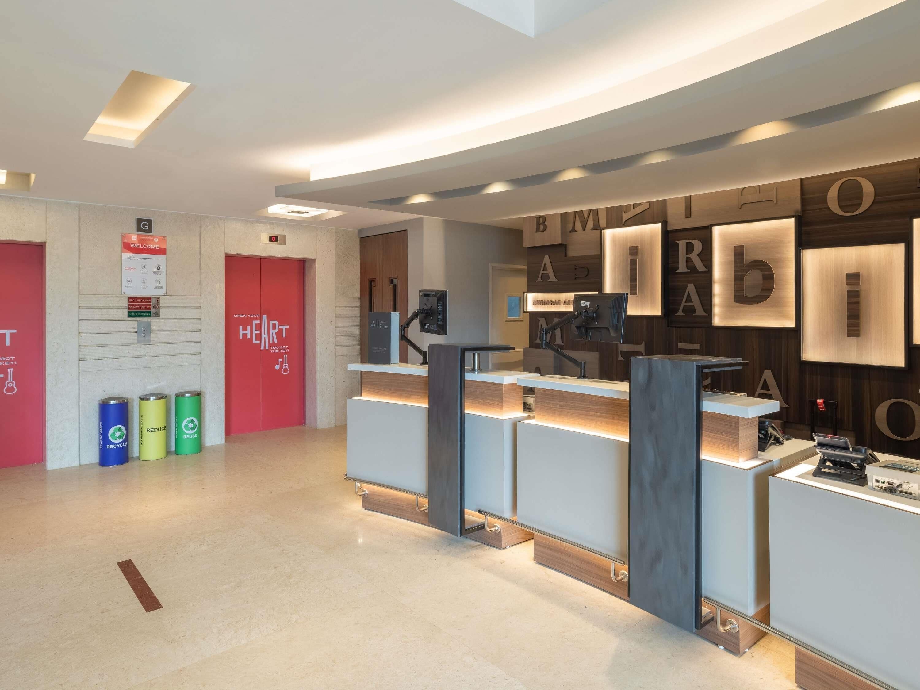 Un área de recepción moderna con una puerta roja, un letrero de bienvenida, contenedores de reciclaje y un escritorio con computadora y teléfono.